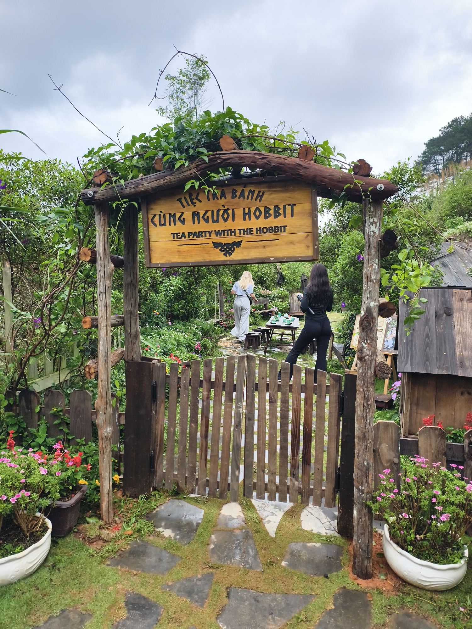 The entrance to a garden or tea party area is shown in the image. A rustic wooden gate with a sign that reads "Tiệc Trà Bánh Cùng Người Hobbit Tea Party With The Hobbit" marks the entrance. The sign is adorned with vines and greenery. Two women are visible beyond the gate, within the garden area. One is wearing a light-colored outfit and is near a table set up with teacups, while the other, closer to the gate, is wearing a black outfit. The ground is covered in grass and stone pavers. Potted flowers are placed on either side of the entrance. A wooden structure is visible on the right. The sky above appears cloudy. The image suggests a garden-themed attraction or tea room, possibly located in Thôn 1, Vietnam.