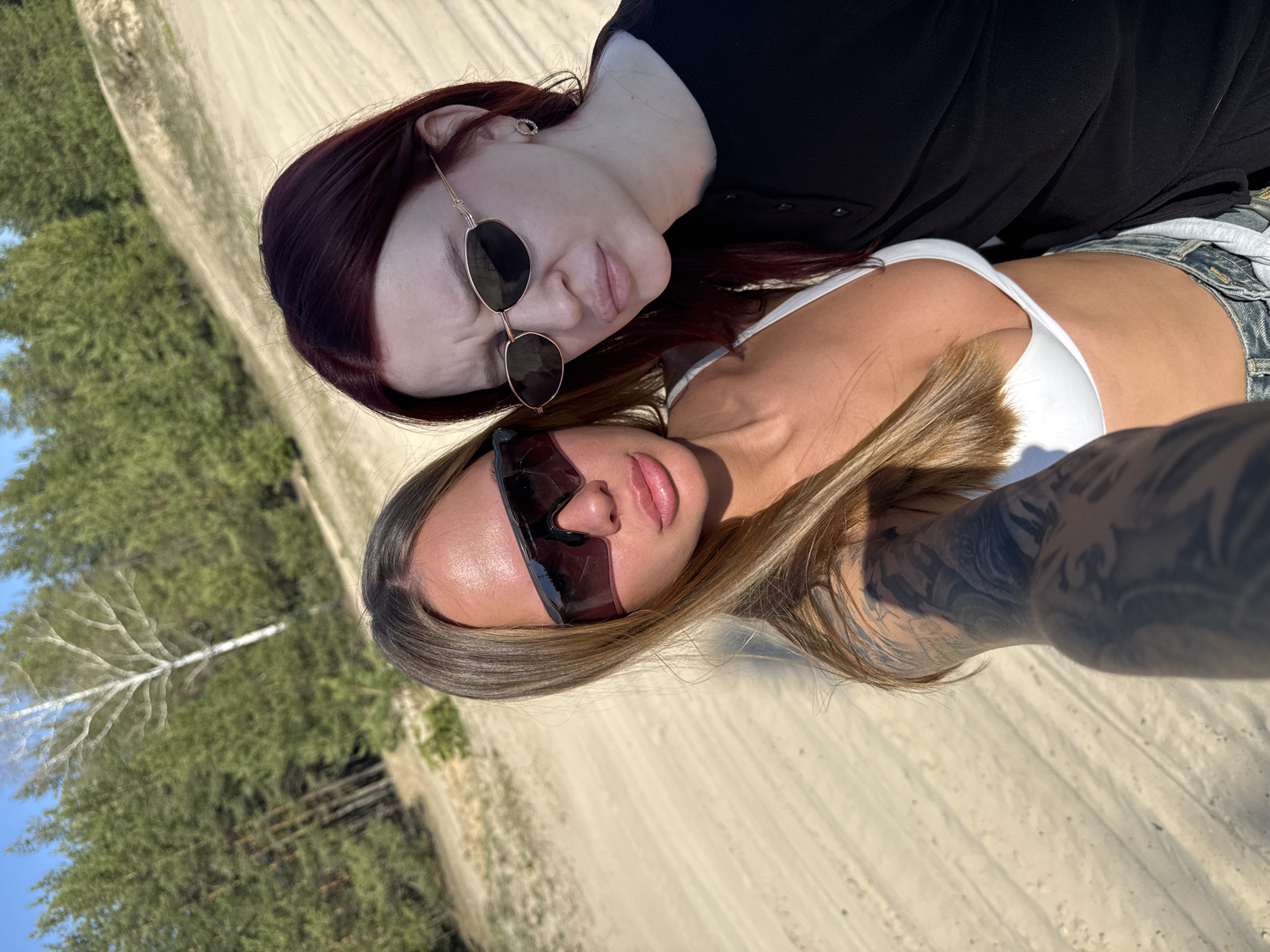 Two women pose for a selfie outdoors on a sunny day. One woman has auburn hair and wears sunglasses and a dark top. The other woman has light brown hair, sunglasses, a white top, denim shorts, and a visible tattoo on her arm. They are close together. The background consists of a sandy or dirt path, trees, and vegetation. The location is likely a park or natural area. There is no evidence suggesting it is in the city of Nizhny Novgorod, Russia.