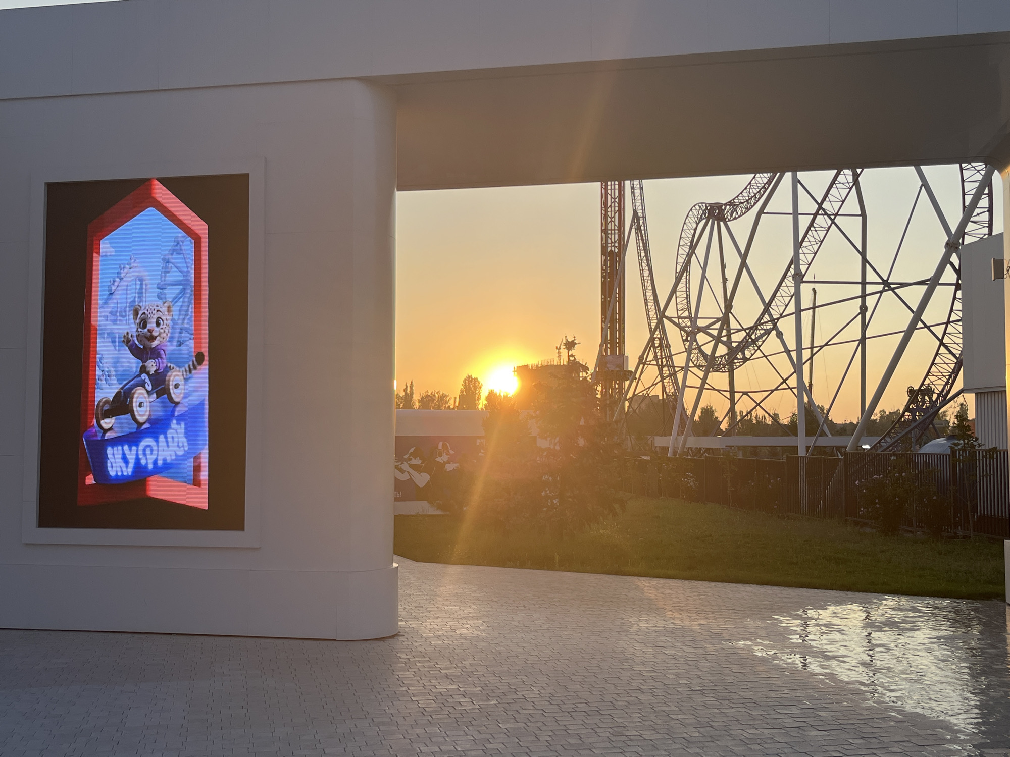 The image shows an amusement park entrance or pathway at sunset. On the left, a large digital screen displays a cartoon cheetah character riding a go-kart, with the words "SKY PARK" in neon blue text below. The screen is framed by a white, modern architectural structure. Through an archway in the structure, the setting sun is visible, casting a warm glow. In the background, a large roller coaster structure dominates the skyline. The ground is paved with light-colored rectangular stones, some of which appear wet, reflecting the light. The environment suggests an outdoor recreational area, likely Bishkek City Park, Kyrgyzstan, given the presence of amusement park rides and a vibrant public space atmosphere.