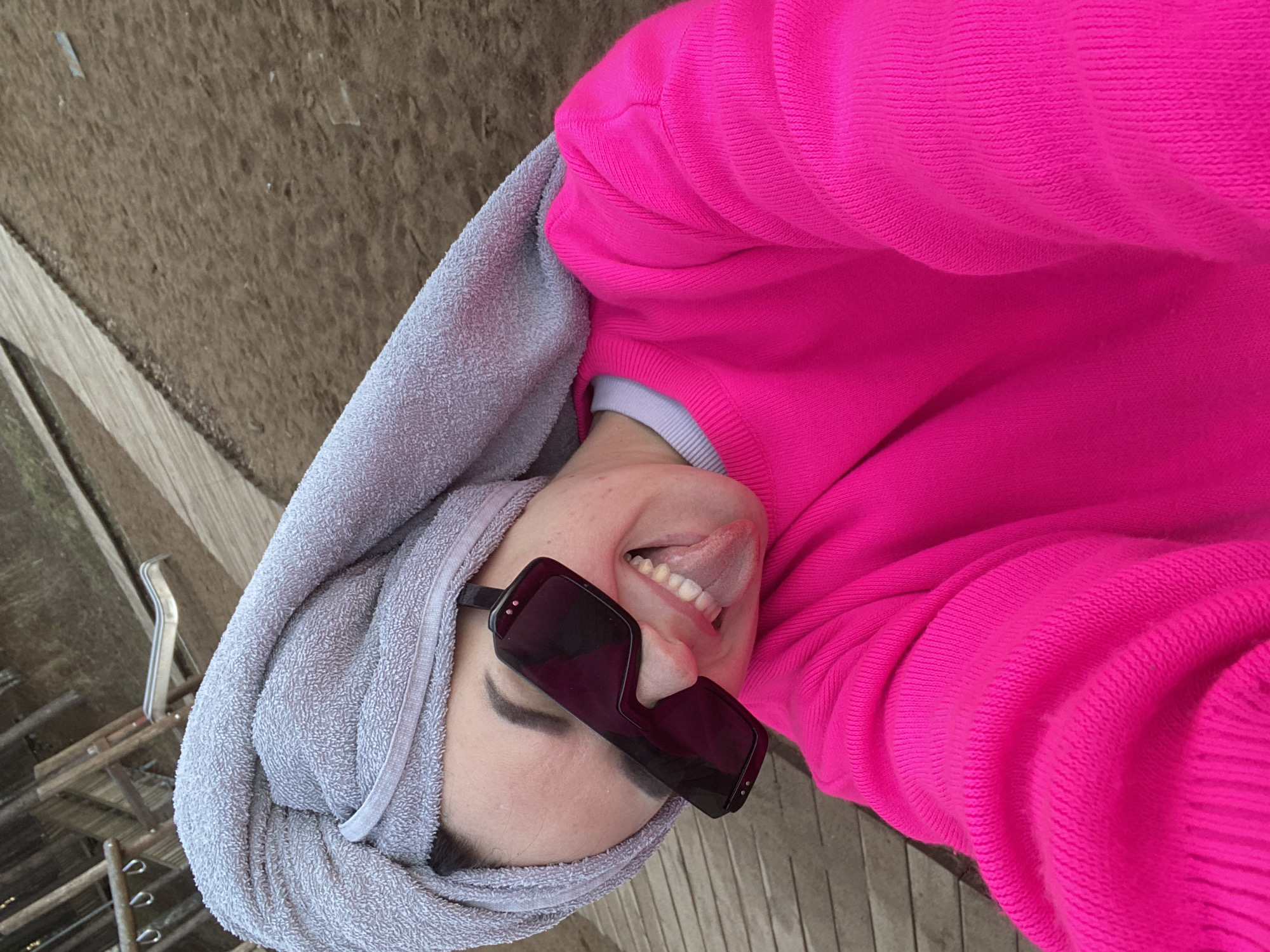 An overhead selfie, rotated counter-clockwise, captures a person (likely a woman) in an outdoor setting in Chaikovsky, Russia. The individual is wearing a vibrant neon pink knitted sweater and oversized dark, rectangular sunglasses. A light grey terrycloth towel is wrapped around their head, suggesting a recent shower or swim. The person is smiling broadly and playfully sticking out their tongue, indicating a cheerful and lighthearted mood. A cuff of a light grey or white undershirt is visible at their right wrist. The background features a textured, earthy ground surface, possibly a path, and on the left, a wooden structure or decking with various metal hooks and elements, suggesting a rustic or casual outdoor environment, possibly near a building or recreational area. The lighting is bright and even, indicating it is daytime with clear weather. No visible text is present in the image.
