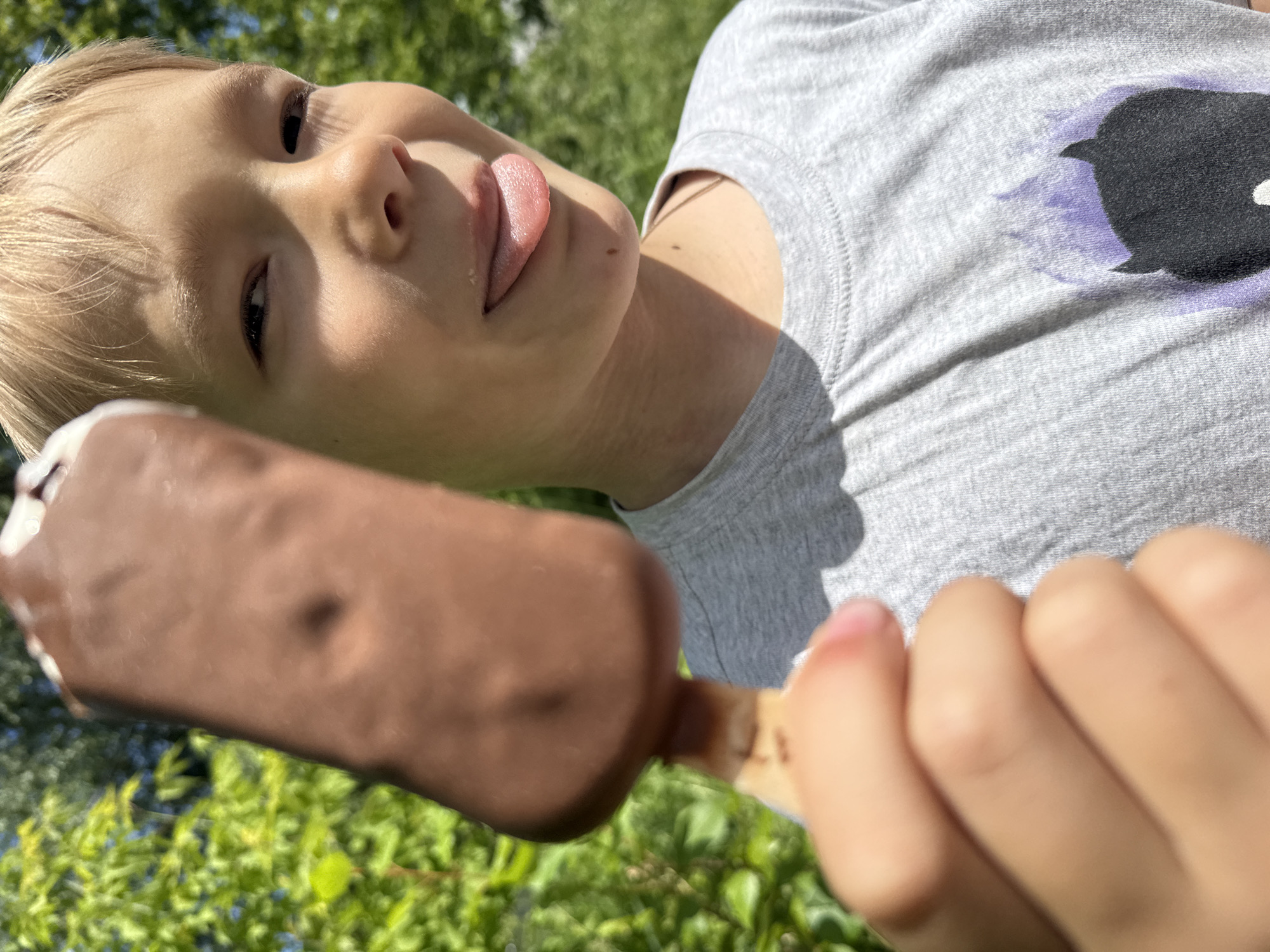In this close-up, low-angle outdoor shot taken in Dmitrov, Russia, a young child with blond hair is the central subject. The child is holding a chocolate-covered ice cream bar on a stick, with their tongue sticking out towards the ice cream. They are wearing a grey t-shirt with a faded black and purple graphic design. The background is filled with lush green foliage and hints of blue sky, suggesting a sunny day, likely in the warmer months. The focus is on the child's face and the ice cream, creating an intimate and playful scene. The child appears to be enjoying their treat, with an expression that conveys a sense of simple pleasure and fun. There is no visible text in the image.