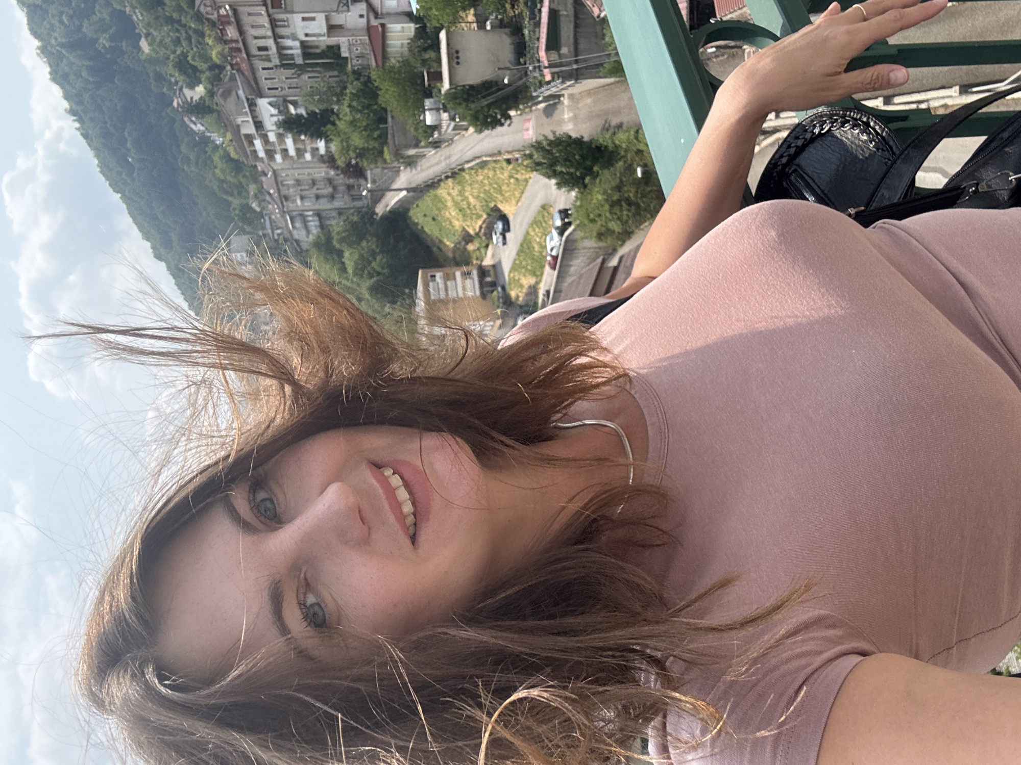 A woman with brown hair and blue eyes is taking a selfie outdoors on a sunny day. She is wearing a pink t-shirt and a silver necklace. Her hair is being blown by the wind. Behind her, there is a view of a hillside town with buildings and trees. The location is Saint-Claude, France.