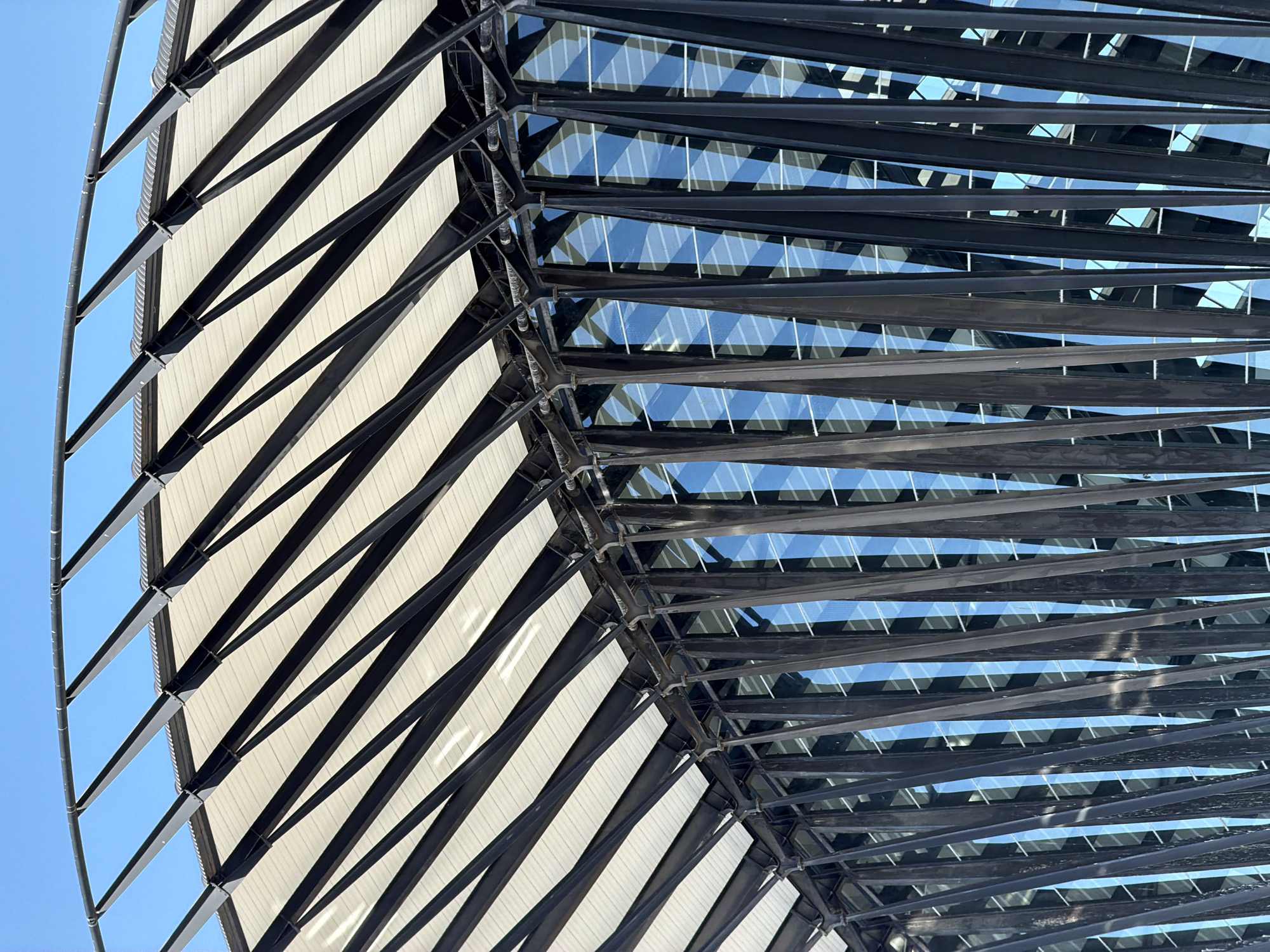 The image shows the exterior of a modern building with a distinctive architectural design. The facade is characterized by a series of angled, dark-colored metal beams that form a rhythmic pattern. These beams are supporting a white, textured canopy that slopes upwards and to the right. Behind the metal beams, large panes of blue glass reflect the clear sky, creating a sense of depth and transparency. The overall impression is one of a contemporary structure with a focus on geometric lines and materials. The scene is set against a bright blue sky, indicating it is daytime and likely sunny. This architectural style is visible at Lyon–Saint-Exupéry Airport (LYS) in Colombier-Saugnieu, France, a location known for its modern airport facilities.