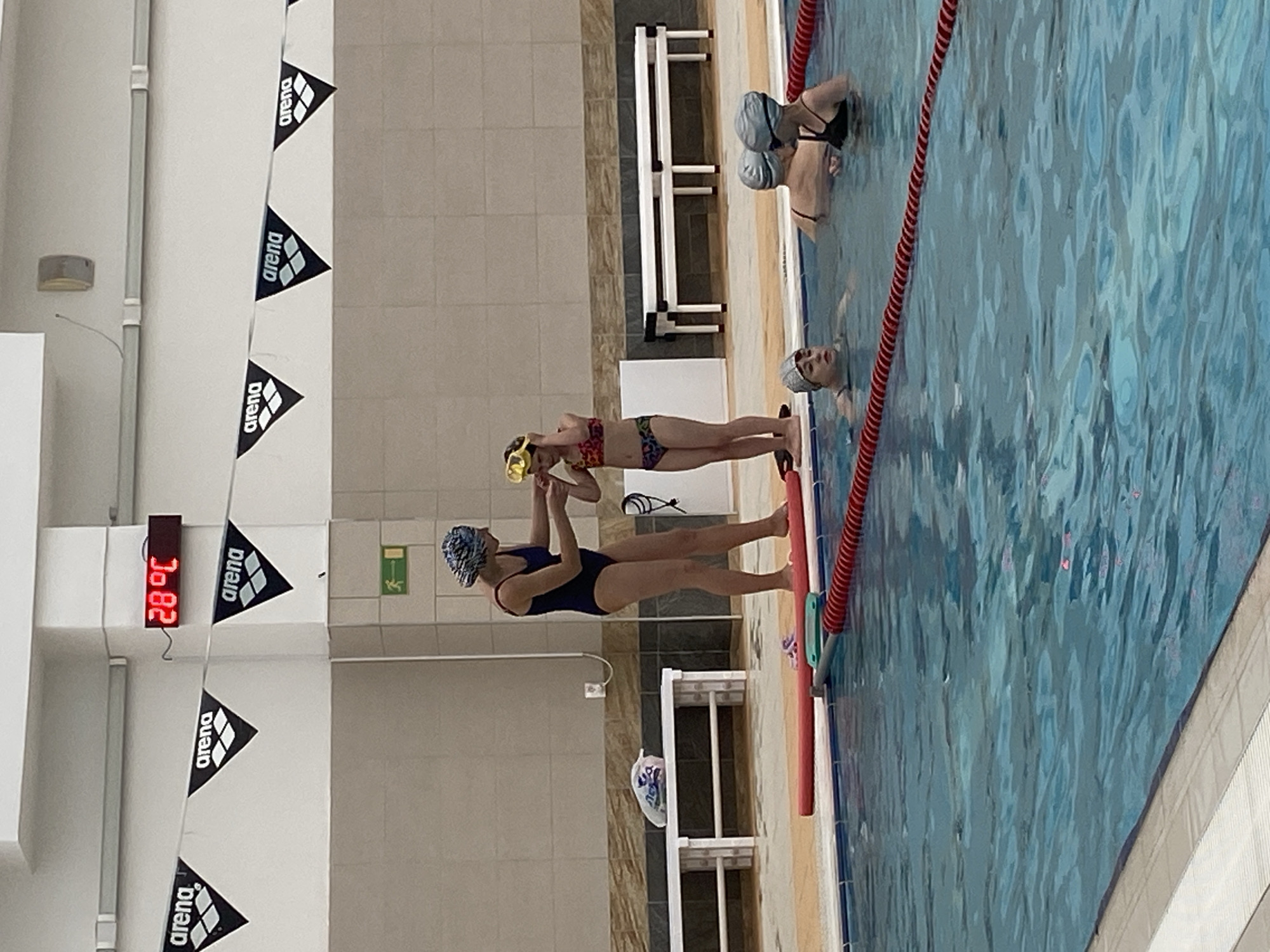 A photo of what appears to be a swimming pool in Yekaterinburg, Russia. A digital thermometer on the wall reads 28 degrees Celsius. There are three people in the water on the right of the frame, wearing swim caps. Two people are standing out of the water to the left of the frame. One adult is wearing a dark blue swimsuit and swim cap, and a child is wearing a patterned bikini and goggles. The adult is holding the child's hands, possibly assisting her or playing with her. "Arena" branded pennants are hung along the wall. Pool lane dividers are visible in the water.