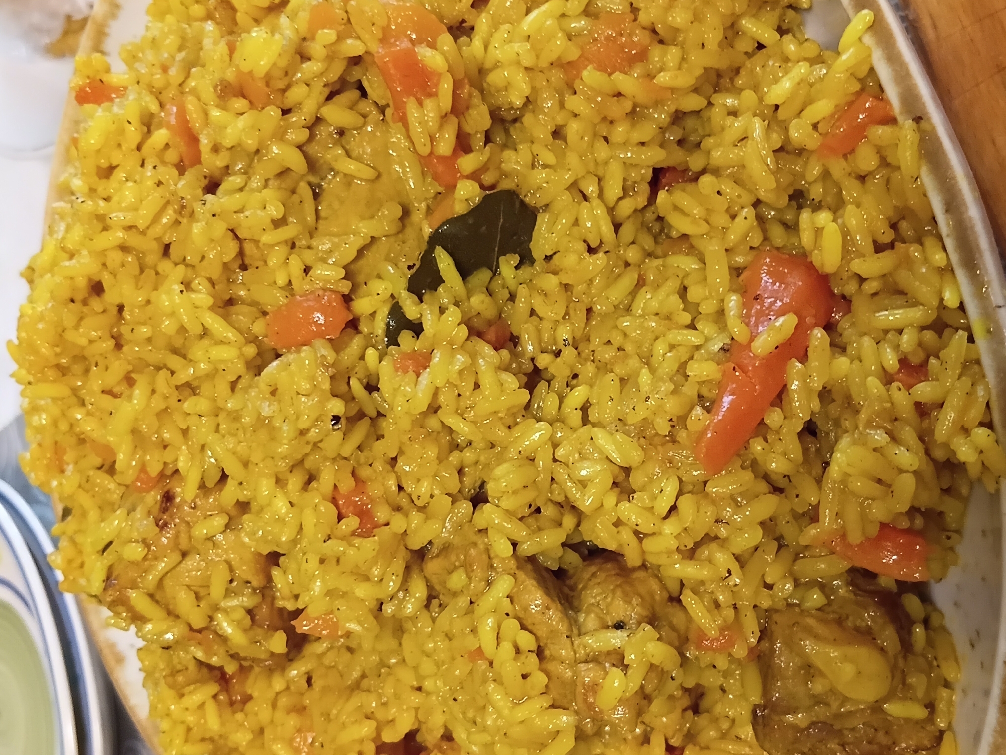 A close-up, overhead shot displays a plate filled with yellow rice mixed with chunks of chicken and pieces of diced tomatoes. The rice appears to be seasoned, with visible specks of black pepper and some green herbs or spices. The dish is served on a beige plate with a brown rim, which is set on a wooden surface. The background shows blurred stacked plates.