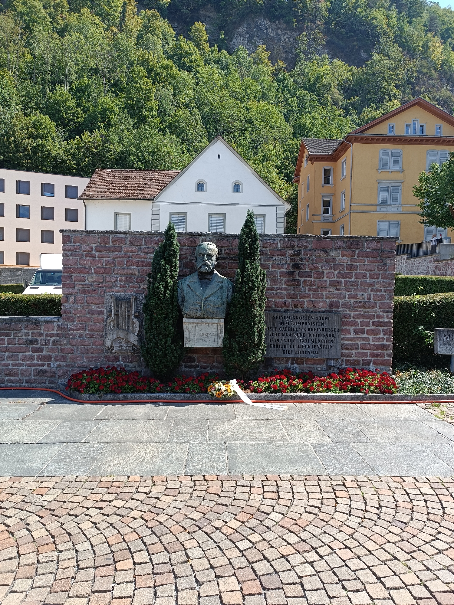 The image depicts a monument in Vaduz, Liechtenstein, dedicated to Josef Gabriel Rheinberger. The monument features a bronze bust of Rheinberger, a composer, mounted on a brick wall. A plaque below the bust is inscribed with text in German, honoring him as a "great son" and "composer." Two cypress trees flank the bust. Red flowers are planted in a bed in front of the wall, and a wreath with a white ribbon lies on the ground. In the background, there are buildings, including a yellow one with light blue shutters and a white one with a red roof. A wooded hillside forms the backdrop. The foreground shows a paved area with interlocking stones. The scene appears to be a public memorial site, bathed in sunlight on a clear day.