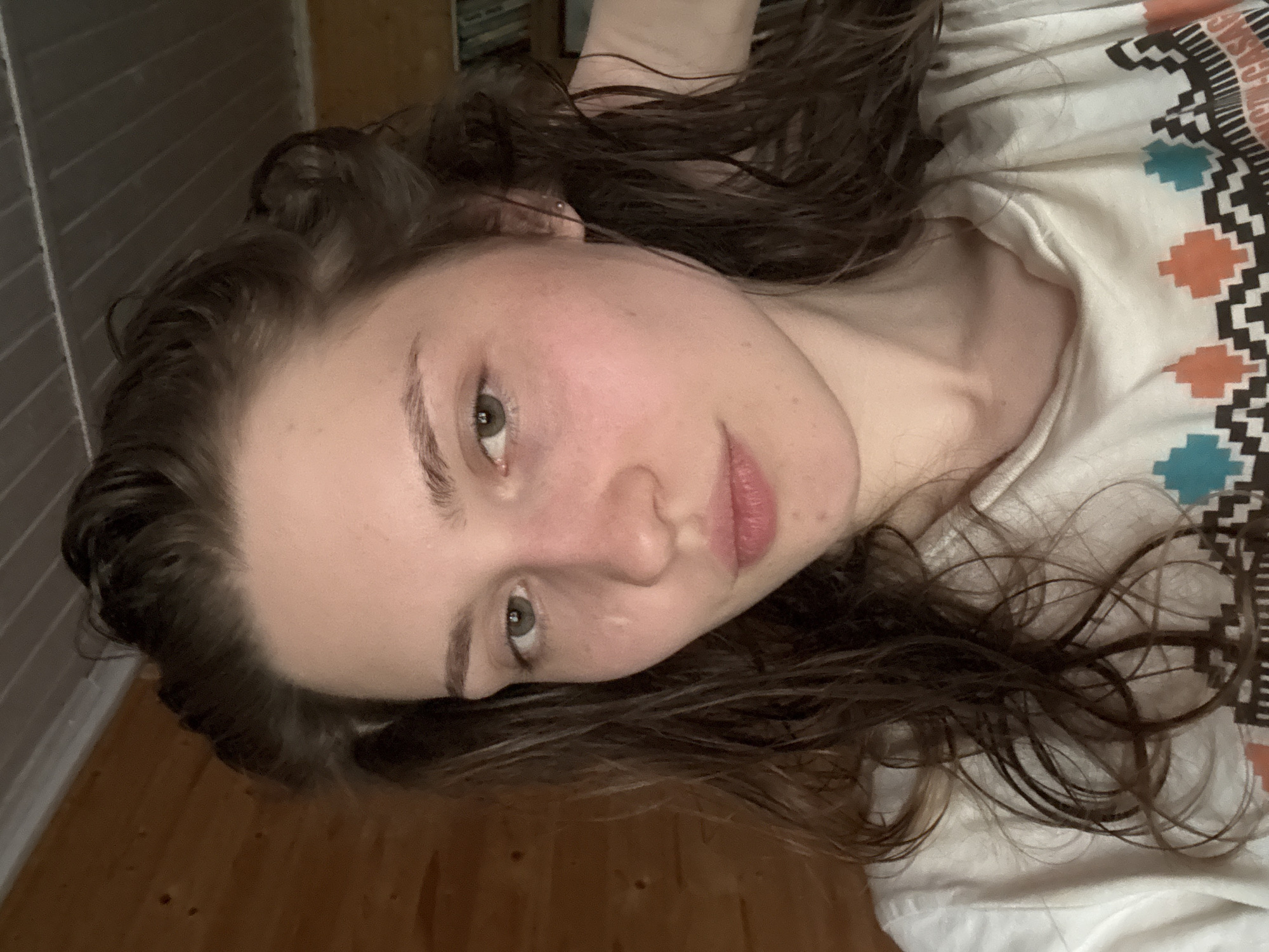 A woman with light skin, brown wavy hair, and green eyes is lying down. She is wearing a white t-shirt with a geometric pattern featuring teal, orange, and black. Part of a wooden floor is visible in the lower left of the frame. The background appears to be a wall with vertical blinds. The location is not definitively identifiable as Ленинградская, Russia, based on the visual information provided.