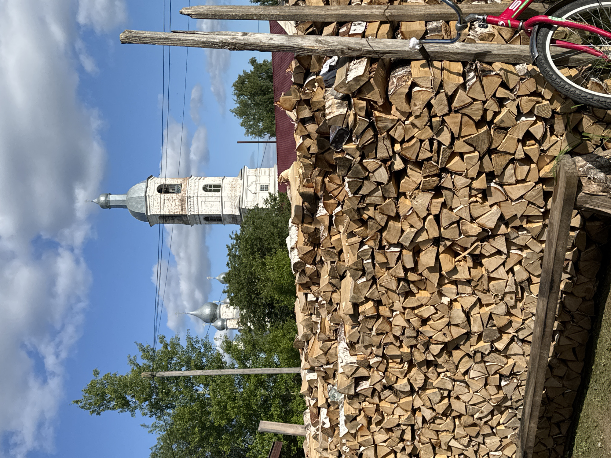 This image depicts a rural or village scene, specified as being in the city of Луптюг, Russia. The foreground is dominated by a meticulously stacked, large pile of split firewood, primarily birch, showing varying shades of light brown and white bark. A rustic, unpainted wooden bench is positioned horizontally at the lower left edge, in front of the firewood. To the right, a partially visible red bicycle with silver fenders and white-walled tires leans against a weathered, rough-hewn wooden post. Another similar wooden post stands further left, behind the woodpile.
Behind the firewood, green deciduous trees obscure the midground. In the background, under a bright blue sky with scattered white cumulus clouds, two distinct church structures are visible. To the right, a tall, white-painted bell tower with multiple arched windows rises, topped by a grey metallic dome and a spire with a cross. To the left of the bell tower, above the treeline, several silver-colored onion domes with crosses are discernible, indicating another church structure. A dark red corrugated metal roof of an unseen building is visible below the bell tower, partially hidden by the woodpile and trees. Thin black power lines traverse the sky horizontally. The scene is illuminated by bright sunlight, casting distinct shadows. No people are present in the image.