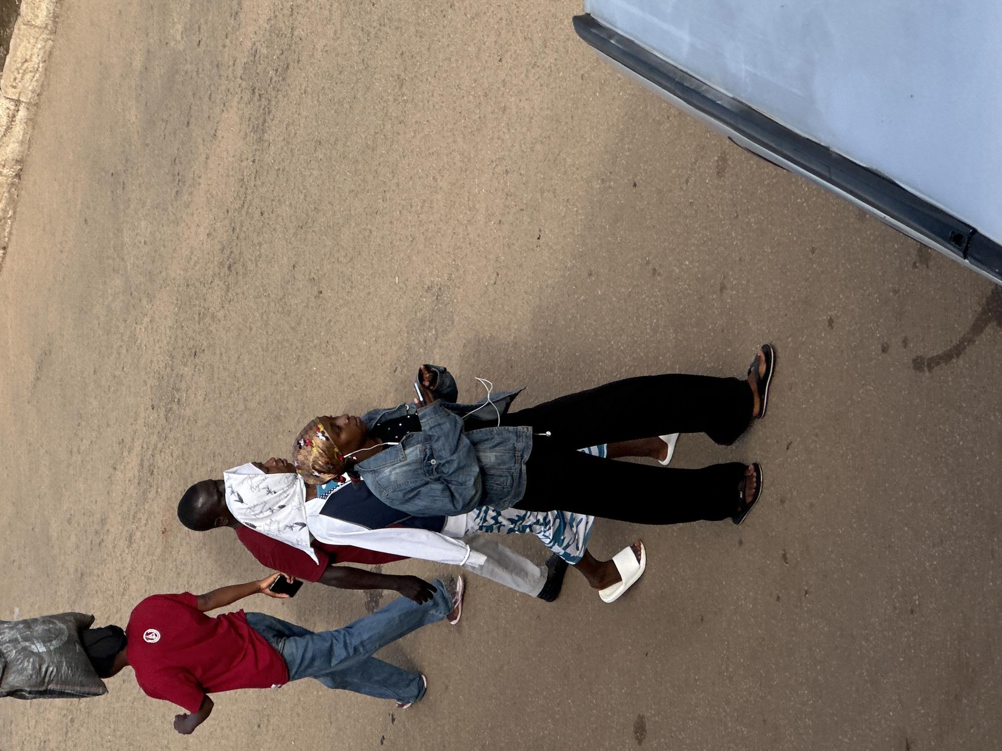 In Bukuru, Nigeria, four individuals walk along a textured, asphalt road. The person in the foreground, wearing a denim jacket and black pants, holds a phone and listens to music through earbuds. Behind them, a person in a white and blue jacket with patterned pants walks. Next to them, another individual in a white headscarf and a red shirt is visible. Leading the group, a person in a maroon t-shirt carries a large, dark sack on their head. The road shows signs of wear and tear, with a curb visible on the left side of the frame. The edge of a white vehicle occupies the lower right corner of the image.