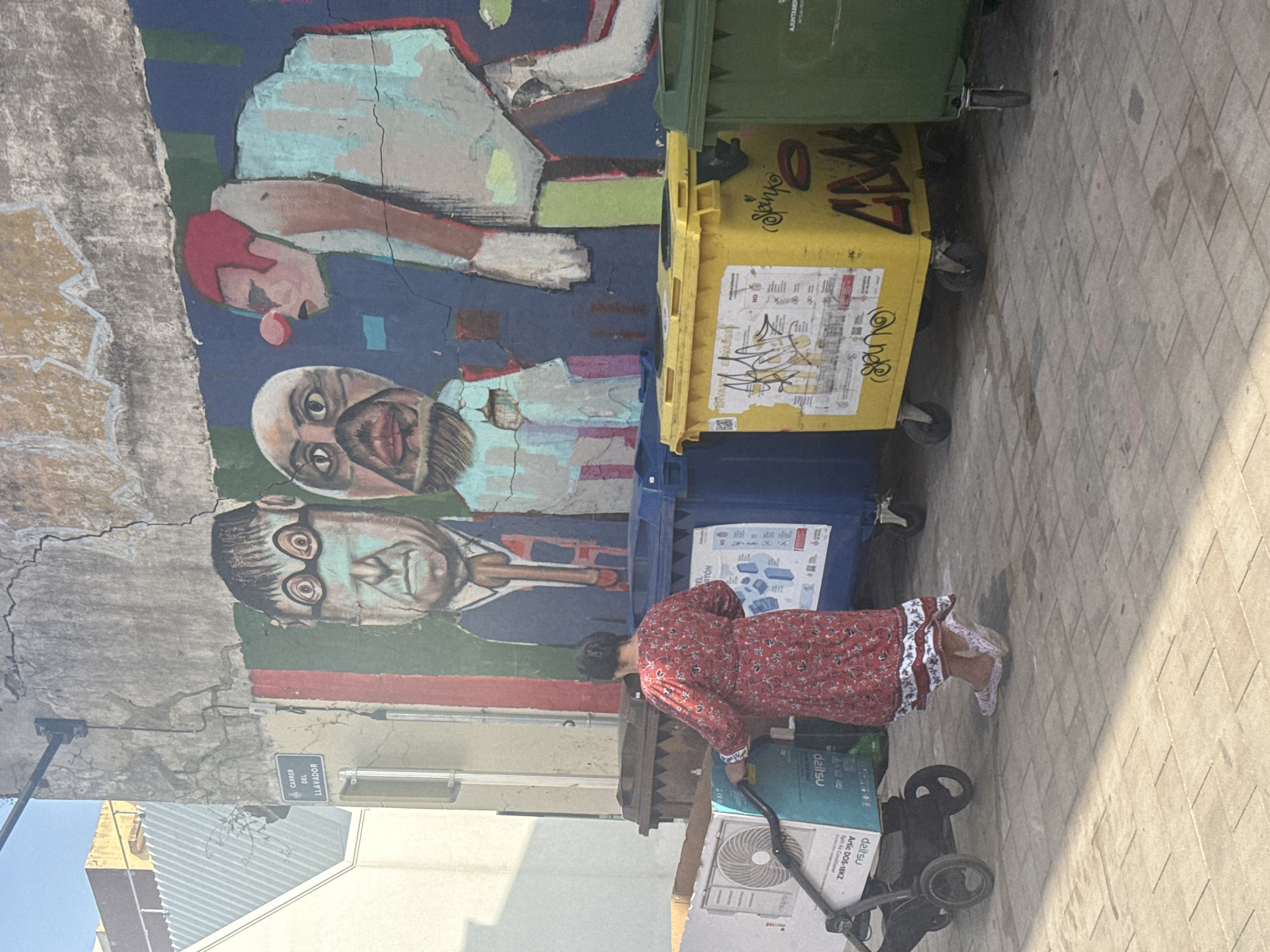 An outdoor street scene in València, Spain, is captured from an elevated perspective, showing a vibrant mural, recycling bins, and a person pushing a cart. The image is rotated 90 degrees counter-clockwise.
Dominating the left side is a large, colorful mural painted on a textured, cracked wall. It features stylized portraits of several men; one with a red-tipped nose, another with a dark beard, and a third with large round glasses. The mural exhibits wear and fading in places.
In front of the mural, three large waste bins are lined up: a green one, a yellow one, and a blue one. These are typical recycling bins, with the green likely for organic waste, the yellow for plastics/cans, and the blue for paper/cardboard. The bins bear graffiti and labels. The green bin has partial text "AJUNTAME" (likely part of "AJUNTAMENT" – City Council) and "توقية". The yellow bin has graffiti including "OHHON", "Janko", "SPUNKo", and a label mentioning "PLASTIC Y ENVASES" (Plastic and Containers). The blue bin has "EL RTON" (likely CARTON) and "PAPEL Y CARTON" (Paper and Cardboard).
A person, likely a woman, is visible on the right, dressed in a red, long-sleeved dress with white floral patterns and lace trim. She is pushing a black hand truck or trolley, which carries a large cardboard box. The box prominently displays "daitsu" and "Artic DDS-18KZ Split Air Conditioner". The ground is paved with square blocks, and sunlight indicates it's daytime, casting shadows.
In the background, on the wall above the bins, a blue street sign reads "CARRER DEL LLAVADOR" (Street of the Washerman), confirming the Spanish location. Other buildings and a bright sky are visible in the distance.