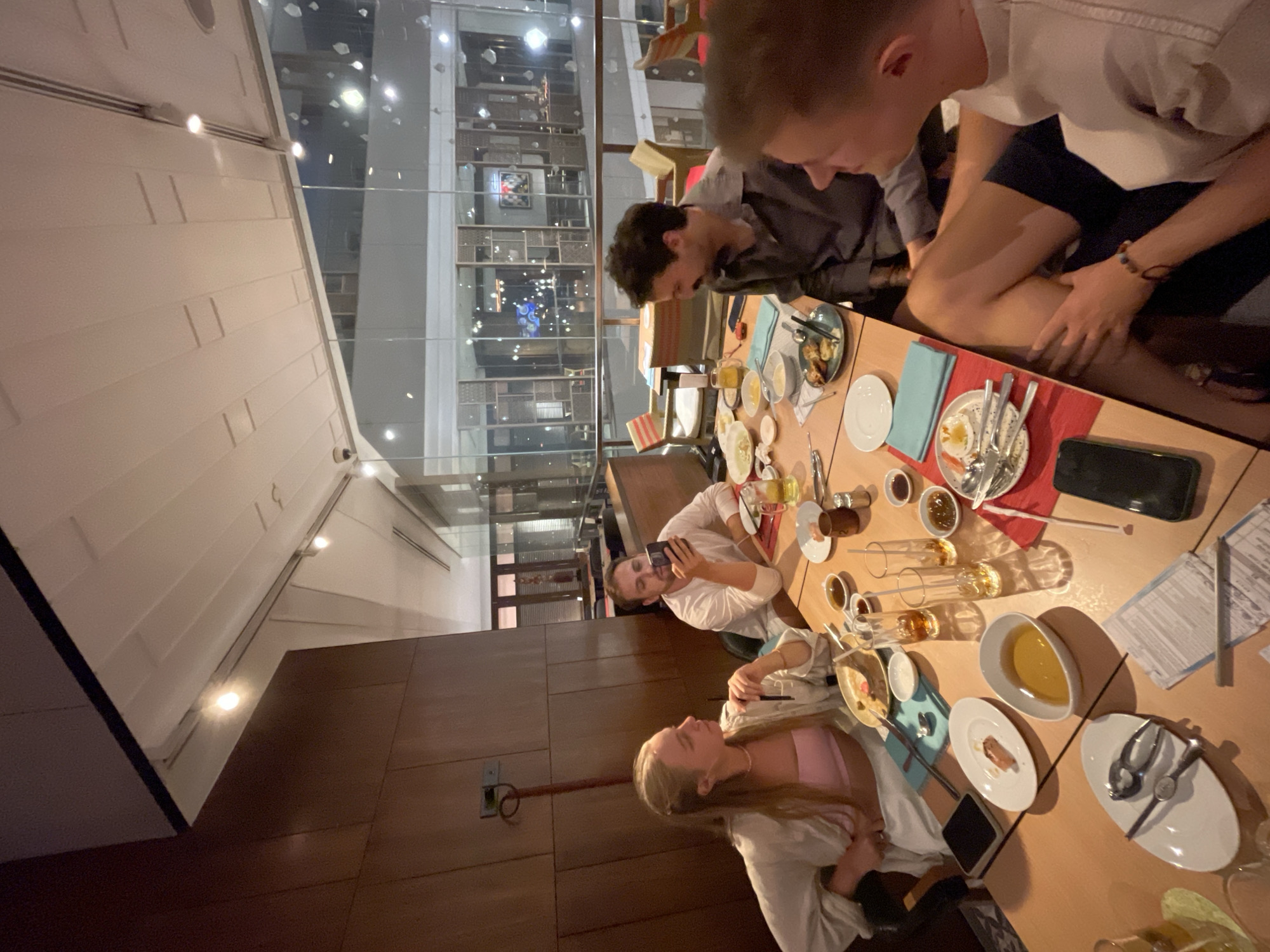 The image shows a group of five people sitting around a table laden with dishes, cutlery, and glassware, indicative of a shared meal. The location appears to be a restaurant in Bangkok, Thailand, judging by the architectural design visible through the large windows, and potentially indoor. People, mostly young adults. One person is using a phone, possibly taking a picture.