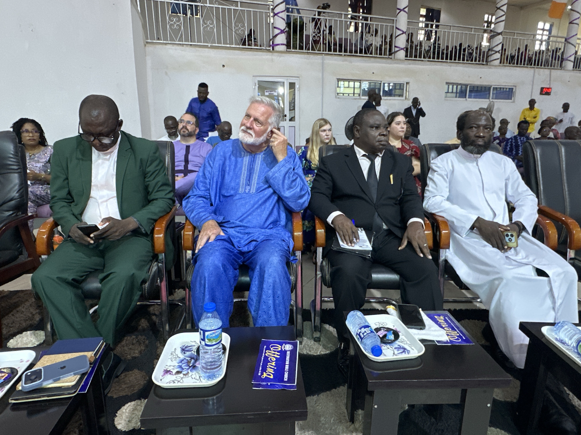 The image shows an indoor gathering, likely a church service, in Television, Nigeria. In the foreground, four men are seated in chairs, facing forward. The leftmost man, wearing a green suit jacket over a white shirt, is looking down at a mobile phone in his hands. Next to him, a man in a bright blue traditional garment, with a white beard, is looking upwards and touching his ear. To his right, a man in a black suit and tie is looking to the right. The fourth man, on the far right, is wearing a white traditional garment and is also looking to the right. Between the men in the foreground and the seating area behind them, there are small tables holding various items including water bottles, trays with floral patterns, and what appears to be a blue booklet labeled "Offering." In the background, more people are seated in rows, and a balcony overlooks the scene, also populated with attendees. The overall environment appears to be a large hall with light-colored walls and a decorative railing on the balcony.