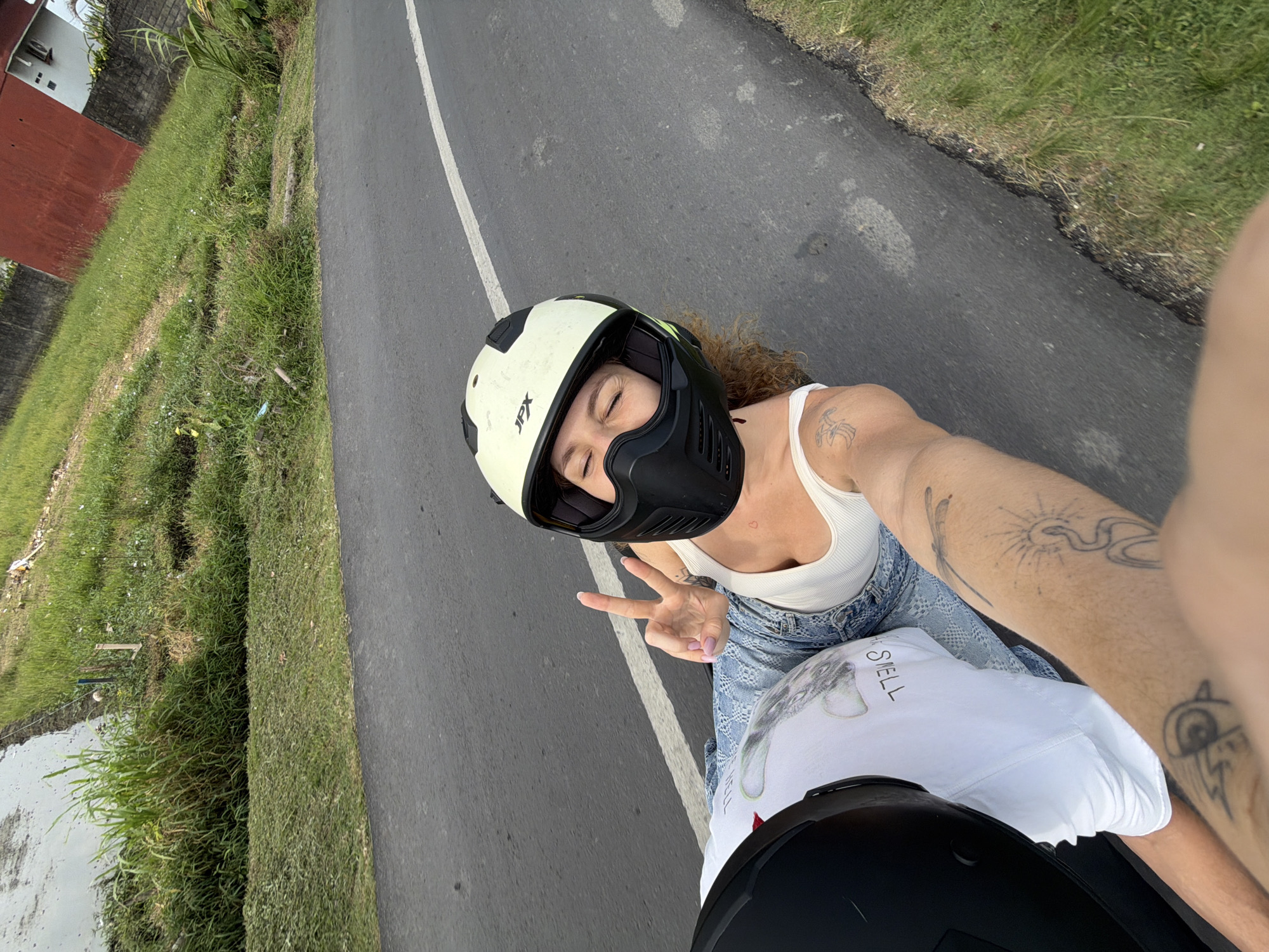 A woman with curly hair, wearing a white tank top, blue jeans, and a white and black helmet, is giving a peace sign while riding on the back of a motorcycle. She is looking at the camera with her eyes closed. The motorcycle is on a paved road with a white line. To the left of the road is a body of water and lush green grass. To the right of the road is more grass and a red building. The image appears to be taken in the city of Mengwi, Indonesia.