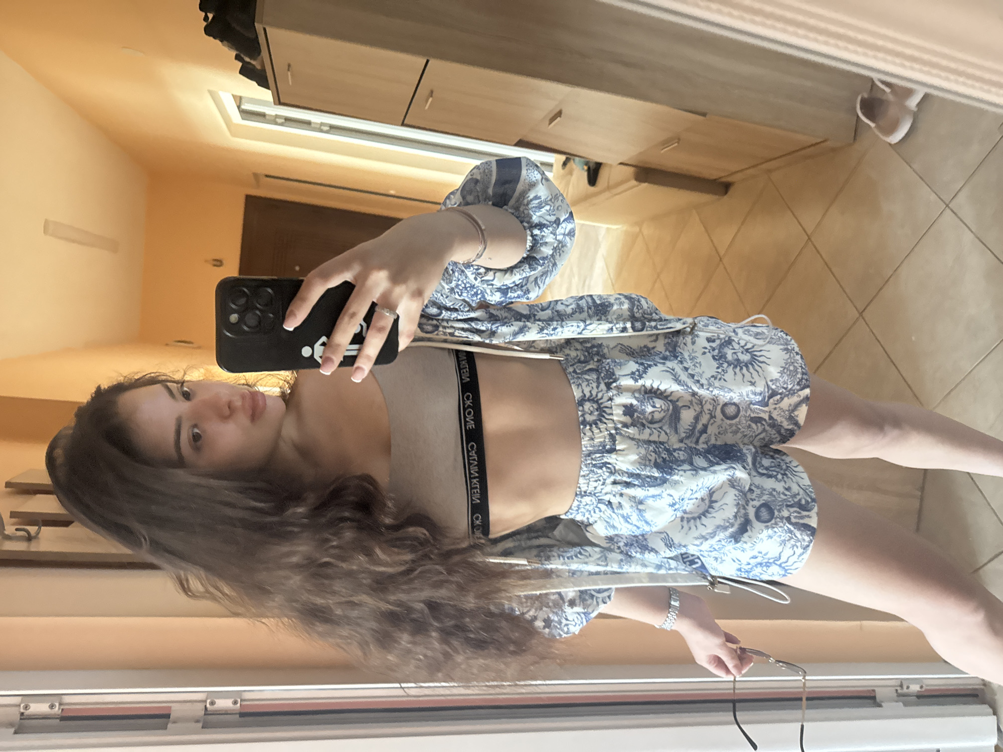 A woman is taking a selfie in front of a mirror. She is wearing a beige sports bra and a blue and white floral patterned shorts set. She is holding a black smartphone in her right hand. Her left hand is holding a pair of glasses. She has long, wavy brown hair. The background shows a hallway with a wardrobe, a door, and a tiled floor. It is not possible to determine the specific location in the city of Plovdiv, Bulgaria, from this image.