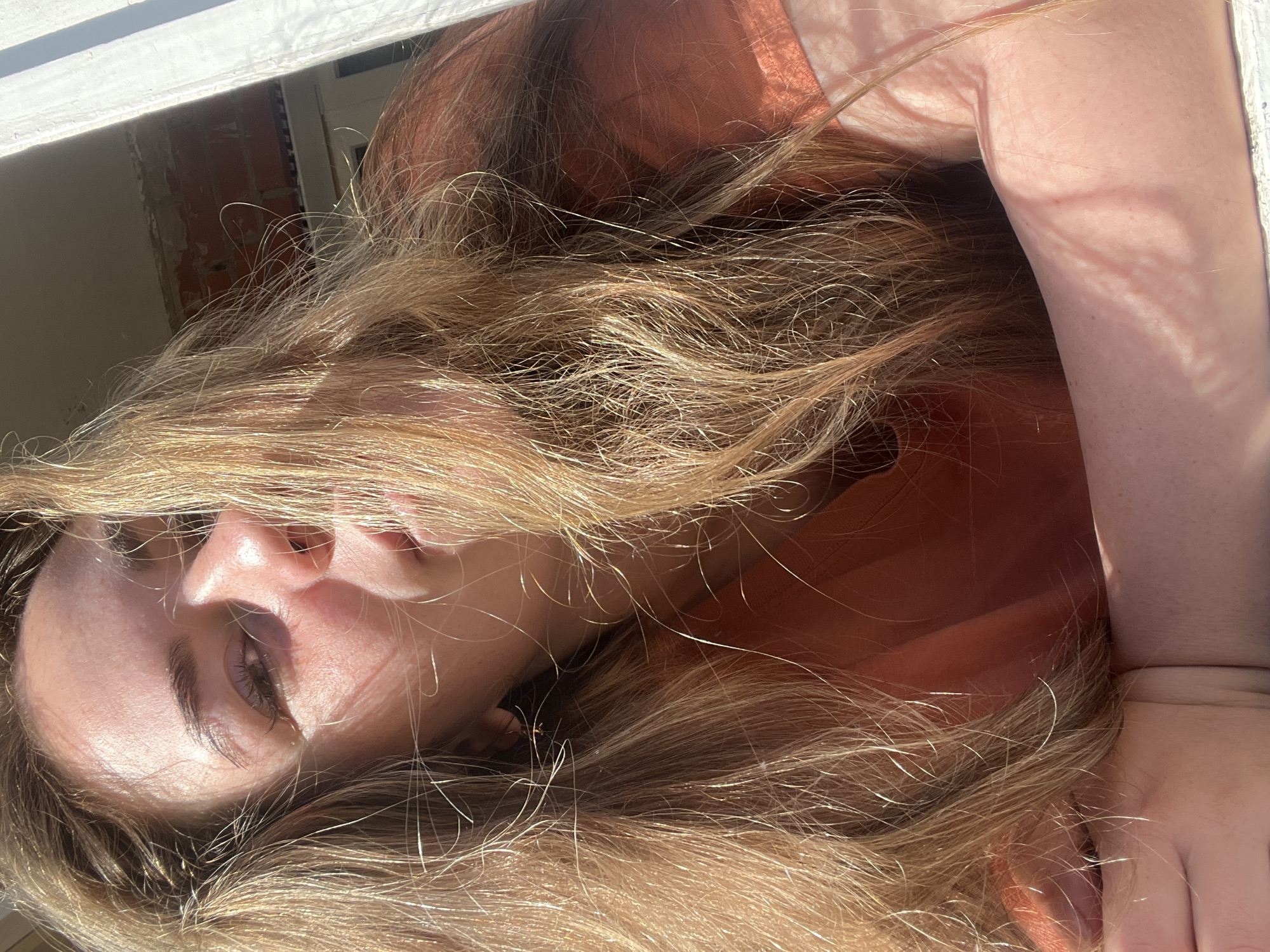A young woman with long, blonde hair is captured in a close-up, outdoor portrait. She is leaning forward, resting her arms on a surface that appears to be a windowsill. The sunlight creates strong highlights and shadows across her face and hair, obscuring some of her features. She is wearing an orange shirt. The background shows a portion of a building under construction, with exposed brick and some roofing materials visible through a window frame. The overall scene suggests a casual, sunlit moment, possibly in a residential area of Chaikovsky, Russia, given the architectural details that are typical of some regions in Russia.