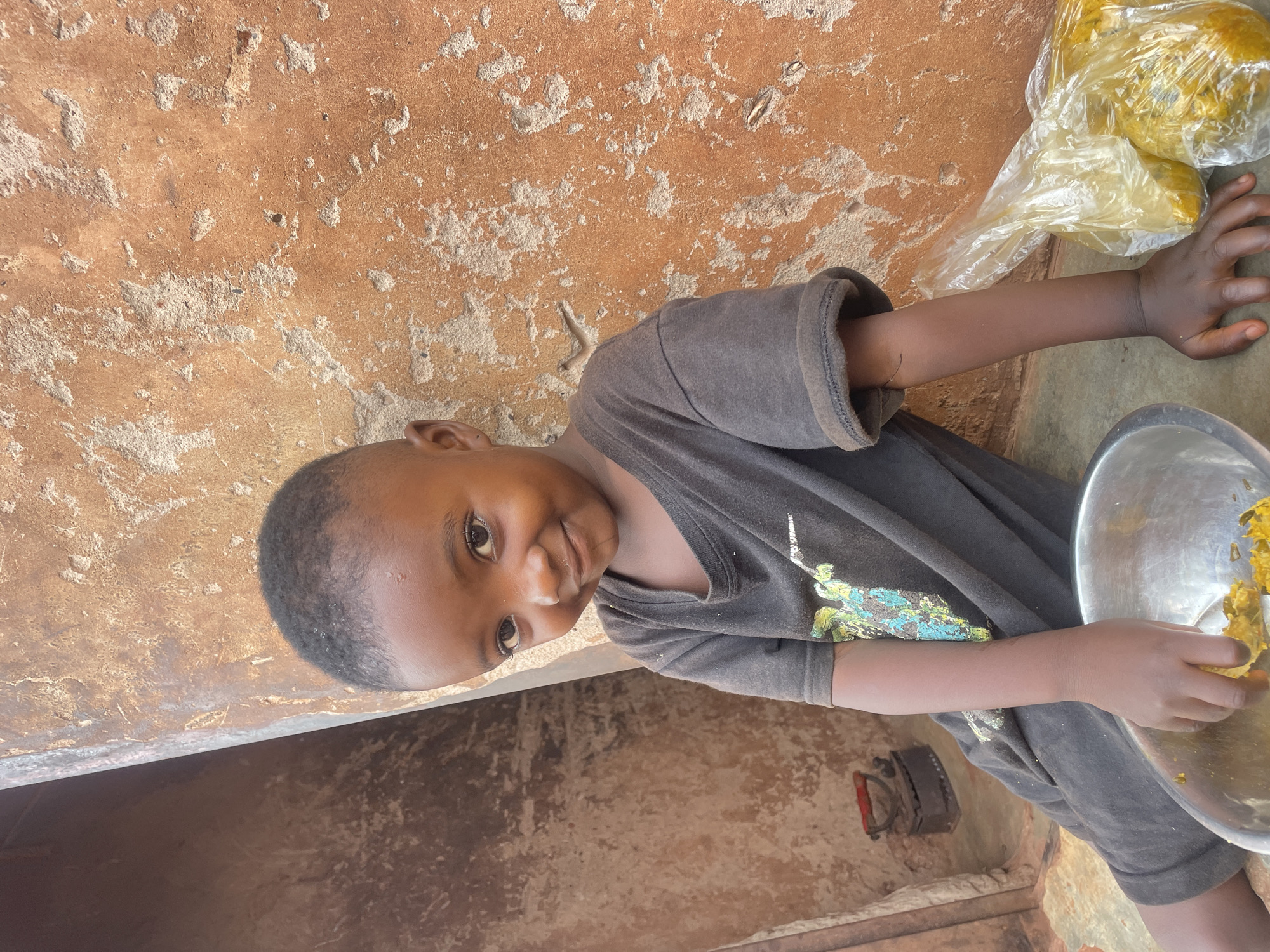 A young child in Ogbe, Nigeria, with dark skin and short hair, stands next to an orange, textured wall. The child wears a grey T-shirt with a colorful design on the front and holds a silver bowl with yellow contents, possibly food. Next to the child’s extended arm is a clear plastic bag filled with yellow items. An old-fashioned metal iron sits near the wall.
