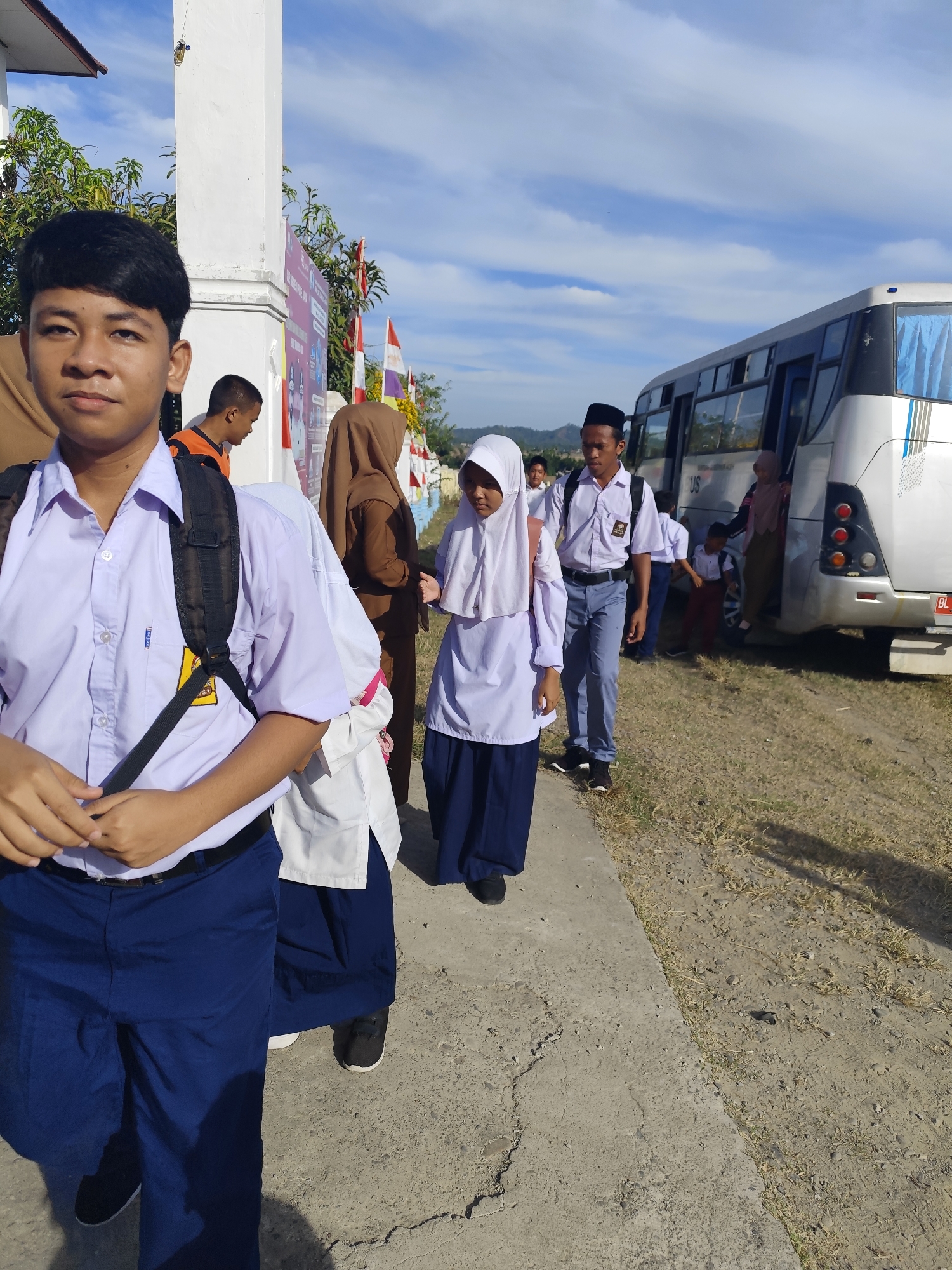 A medium shot captures a group of students in uniform walking along a concrete path next to a road. The student in the foreground, a male with short black hair, wears a light purple button-up shirt, dark blue pants, and a black backpack. He faces the camera with a neutral expression. Behind him, several other students are visible. A female student in a white hijab and uniform walks to his left. To the right of the foreground student, a male student in a white uniform and black peci hat walks, followed by other students. A white bus with blue-tinted windows is parked on the right side of the frame, with some students entering or exiting it. The environment appears to be outdoors with dry grass and dirt on the side of the road, and a blue sky with scattered clouds above. Flags are visible in the background, attached to a white pillar.