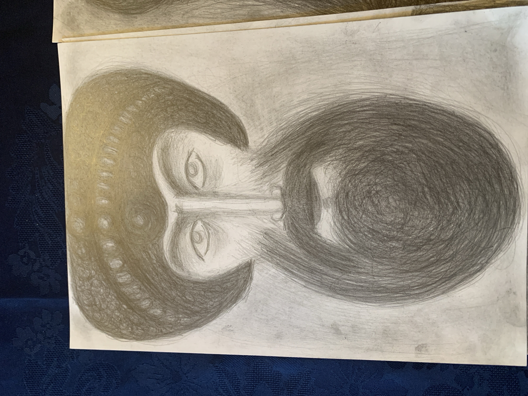 A pencil drawing depicts a man's face from the shoulders up. He wears a turban or hat with textured bands. His eyes are large and almond-shaped, with prominent eyebrows. A long, bushy beard fills the lower half of the drawing, rendered with dense, swirling pencil strokes. The drawing is on a piece of paper that appears to be slightly yellowed and is placed on a dark blue, textured fabric.