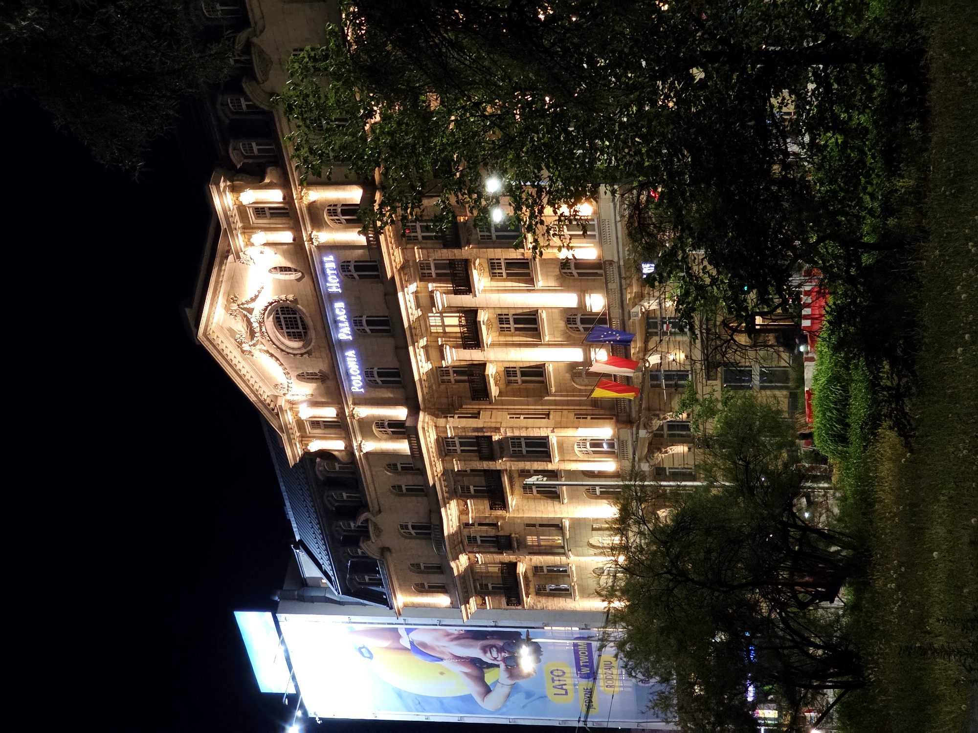 The image shows the Polonia Palace Hotel in Warsaw, Poland, at night. The hotel is illuminated by warm lights, highlighting its architectural details. To the left of the hotel, a large billboard displays an advertisement featuring a smiling woman with a yellow inflatable. The billboard text includes the word "LATO" (Summer in Polish). In front of the hotel, there are trees and greenery, with a patch of grass and some scattered white flowers in the foreground. The sky is dark, indicating nighttime. Flags, including the Polish flag and the European Union flag, are visible on the hotel building.
