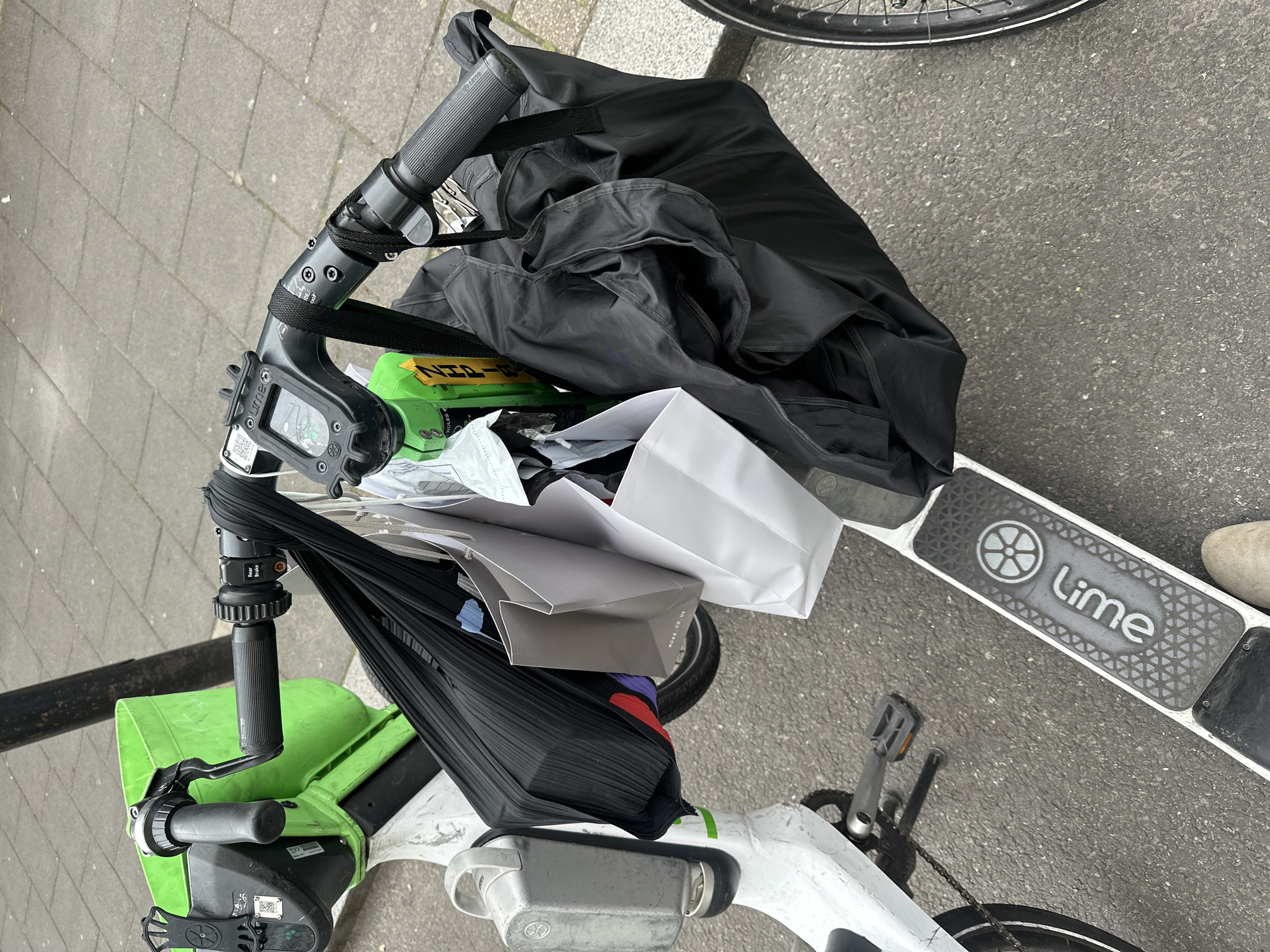 A white and green Lime electric bicycle is parked outdoors on an asphalt road adjacent to a grey concrete paved sidewalk. The handlebars and front basket are heavily laden with various shopping bags and items. Prominently visible are a large black pleated fabric bag, a grey paper shopping bag with "MOSS.CO.UK" printed on its base, and several other white paper and black fabric bags. Tucked among the bags on the right side is a bright green bottle or container with partially visible yellow text reading "ZHP-1B" and "RULES."
The e-bike features black handlebars with ribbed grips, front and rear brake levers (the right one labeled "Rear Brake"), a central digital display unit, and a phone mount on the left side of the stem. A black strap is wrapped around the center of the handlebars, securing some of the bags. The white frame shows signs of use, and the "Lime" logo is clearly visible on the white footboard platform. The left pedal and drive chain are visible. In the bottom right corner, a person's right foot, clad in a light tan suede-like shoe, is positioned on the ground near the e-bike's footboard. The ground transitions from dark asphalt to light grey paving slabs, with a dark vertical pole visible in the background. The scene is illuminated by an overcast sky, indicating a public street in London, United Kingdom.