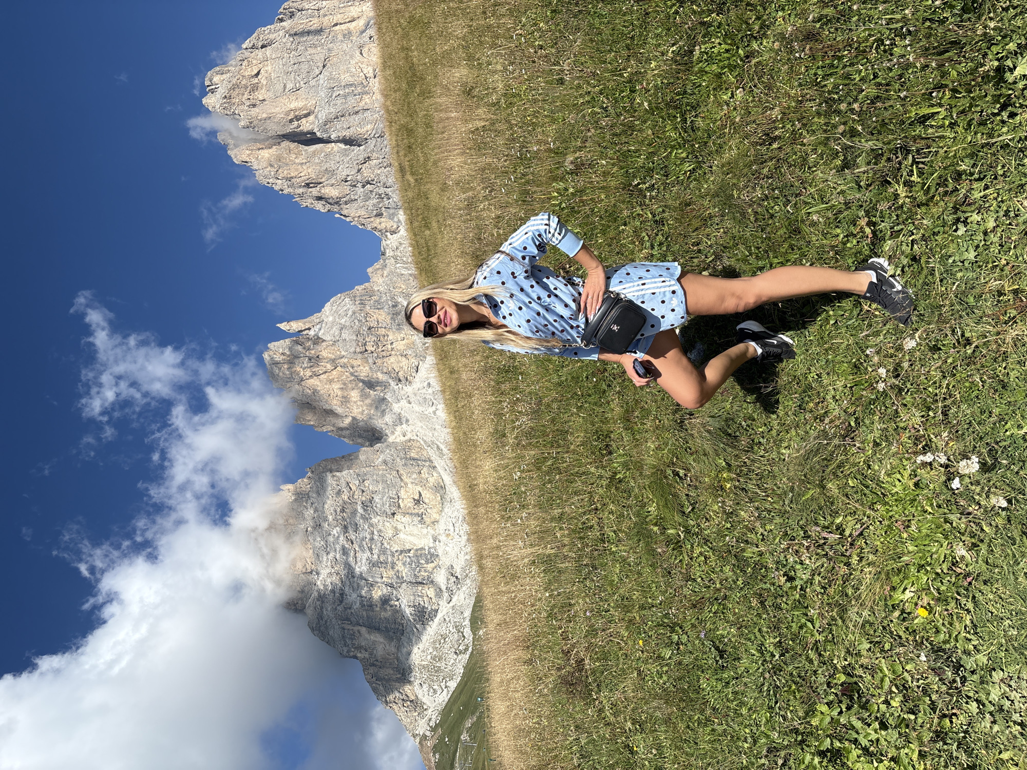 A blonde woman stands on a grassy hillside in front of three large, rocky mountains under a bright blue sky with scattered clouds. The woman is wearing a light blue dress with black polka dots, a black fanny pack, sunglasses, and black sneakers. Her left hand is on her hip, and her right hand is resting on her left thigh. The mountains in the background appear to be partially covered in clouds. The grassy slope is green in the foreground and transitions to dry, golden grass further up the hill. The location is identified as the Dolomites, near Canazei, Italy.