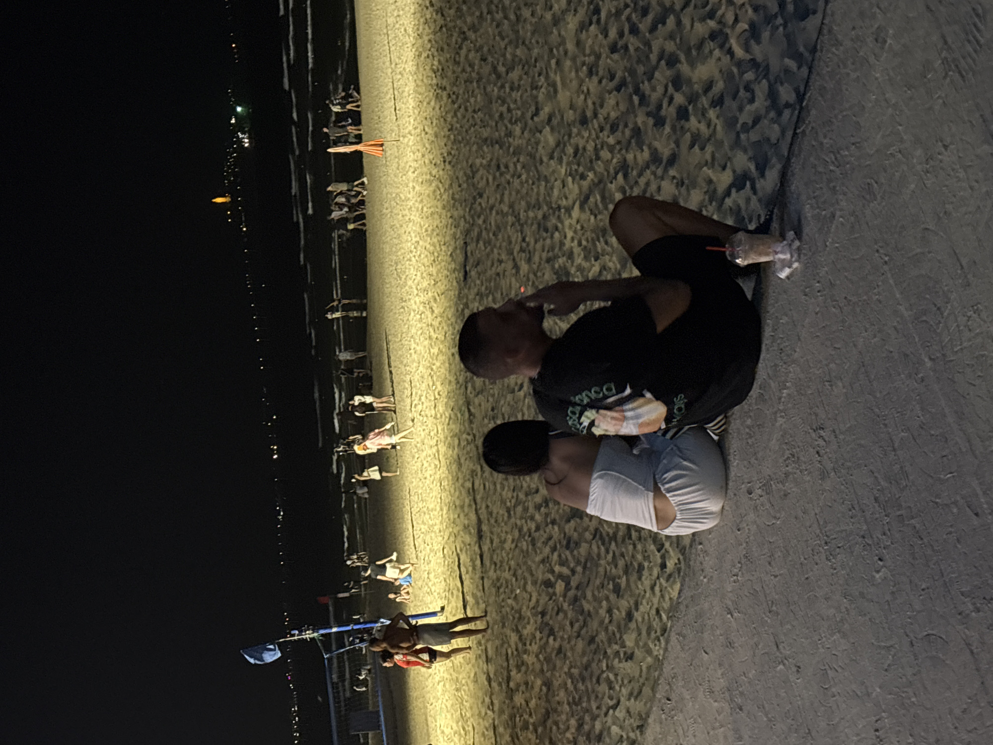 Captured at night, the scene unfolds on a beach in Đà Nẵng, Vietnam. A man and a woman are seated closely together on the sand, near a concrete walkway. The man is wearing a black shirt and shorts. He holds a drink in a plastic cup with a straw. The woman is wearing a white top. In the background, a beach volleyball net is visible, along with numerous individuals scattered along the beach. Distant city lights illuminate the horizon. The beach is covered in sand.