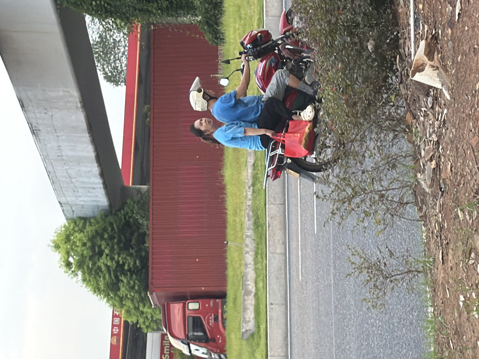 The image depicts a street scene in Jiangjin, China, likely a suburban or industrial area given the presence of a large red shipping container and what appears to be a gas station canopy in the background. Two individuals, a woman and a man, are riding on a red motorcycle. The woman is seated behind the man, wearing a blue t-shirt and black pants, and is carrying a red tote bag. The man is driving the motorcycle, wearing a blue t-shirt, grey shorts, and a white helmet. A red truck is partially visible to the left of the frame, next to the shipping container. The motorcycle is on a paved road with white lane markings, and there are green grassy areas and vegetation on either side of the road. An elevated concrete structure, possibly part of an overpass or bridge, is visible in the upper portion of the image. The sky is overcast.