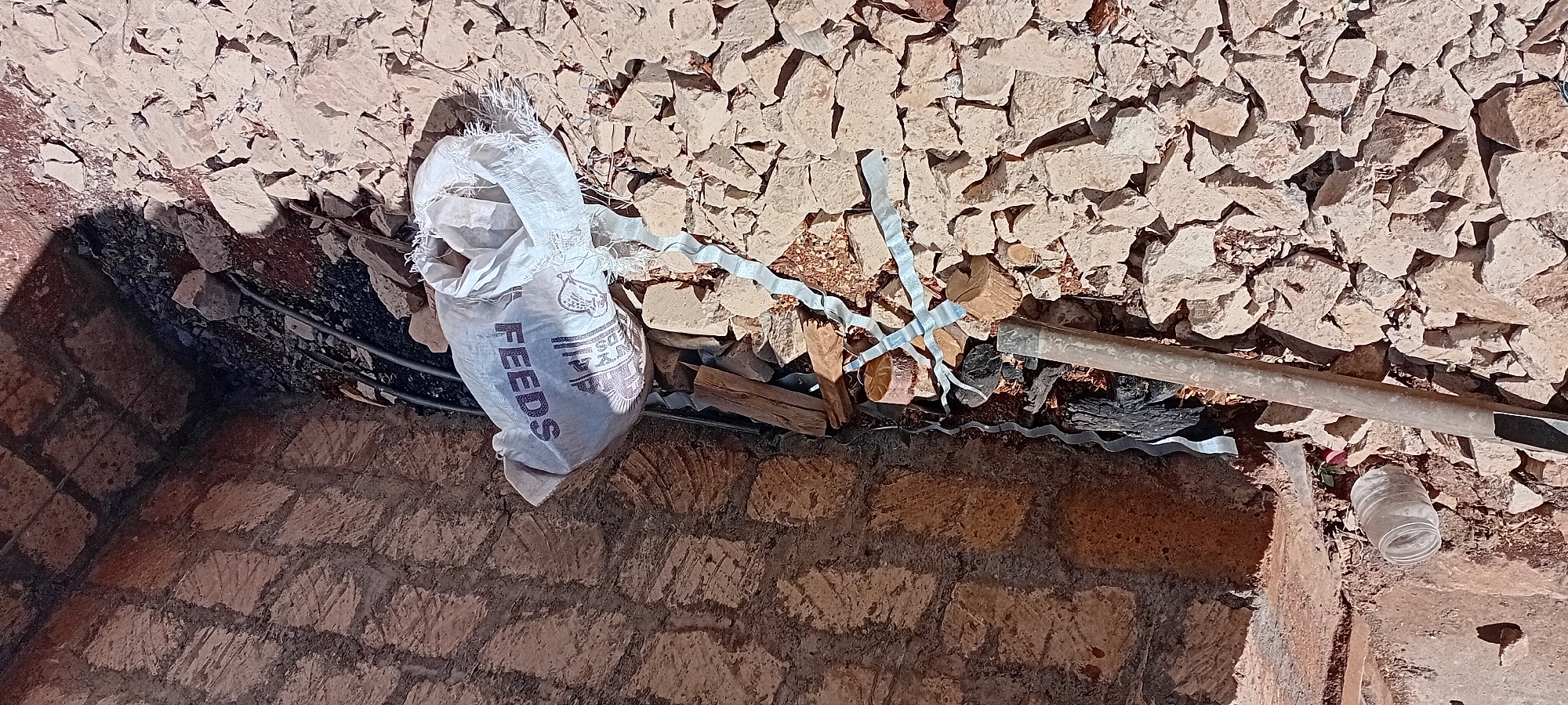 A scene of debris and construction materials is shown in Kijabe Ward, Kenya. The ground is covered with numerous small, rough-hewn stones and dirt. In the foreground, a silver-colored pipe runs horizontally across the frame. To the right of the pipe, there is a white plastic bag with the word "FEEDS" printed on it in large, dark lettering, along with some other unreadable text and graphics. The bag appears to be partially filled and is open at the top, with frayed edges. Scattered around the bag are pieces of wood and some dark, charred material, possibly remnants of a fire. To the left of the pipe, a clear glass jar lies on its side amidst the stones. The background consists of a rough, stone-built wall, showing evidence of construction or age. The lighting suggests it is daytime, with shadows cast by the wall.
