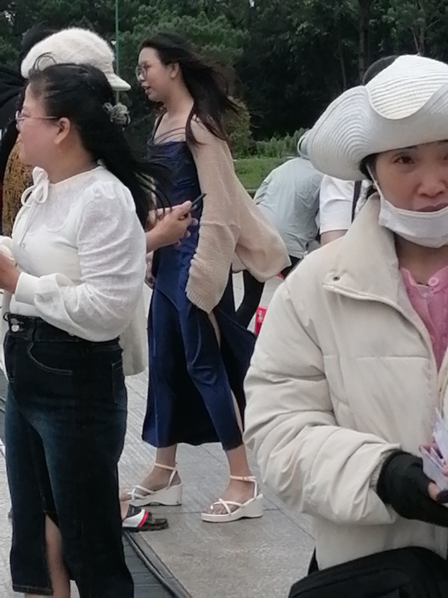 The image shows a group of people outdoors, likely in a public space in Da Lat, Vietnam. In the foreground, a woman wearing a white quilted jacket and a large white hat with a brim is visible from the chest up. She is also wearing a white face mask. To her left, a woman in a white long-sleeved shirt with a ribbon tie at the neck and dark jeans is partially visible. Behind her, another woman in a dark blue dress and a light-colored cardigan is in profile, holding a dark object, possibly a phone, in her hand. The background consists of trees and foliage, suggesting a park or a natural setting. The ground appears to be paved. The overall lighting suggests it is daytime.