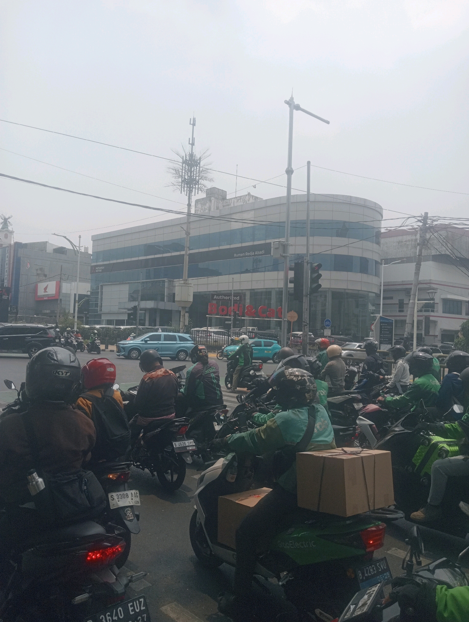 This is an outdoor, eye-level, full-shot photograph of a congested street in South Jakarta, Indonesia, on a hazy day. The foreground is dominated by a large number of motorcycles and their riders, many wearing green jackets and helmets, suggesting they are likely delivery drivers. The motorcycles are stopped, indicating traffic or a red light. Some riders are looking forward, while others are looking around.
In the midground, there are more vehicles, including cars, also stopped in traffic. The buildings in the background are modern, with one prominent building featuring a curved glass facade. Signage on the buildings indicates commercial establishments, including "Bumen Reja Abadi" and "Authorized Hi & Cat". Traffic lights and utility poles are visible throughout the scene. The overall atmosphere is one of urban hustle and bustle, with a significant presence of motorcycles being used for transportation and delivery services.