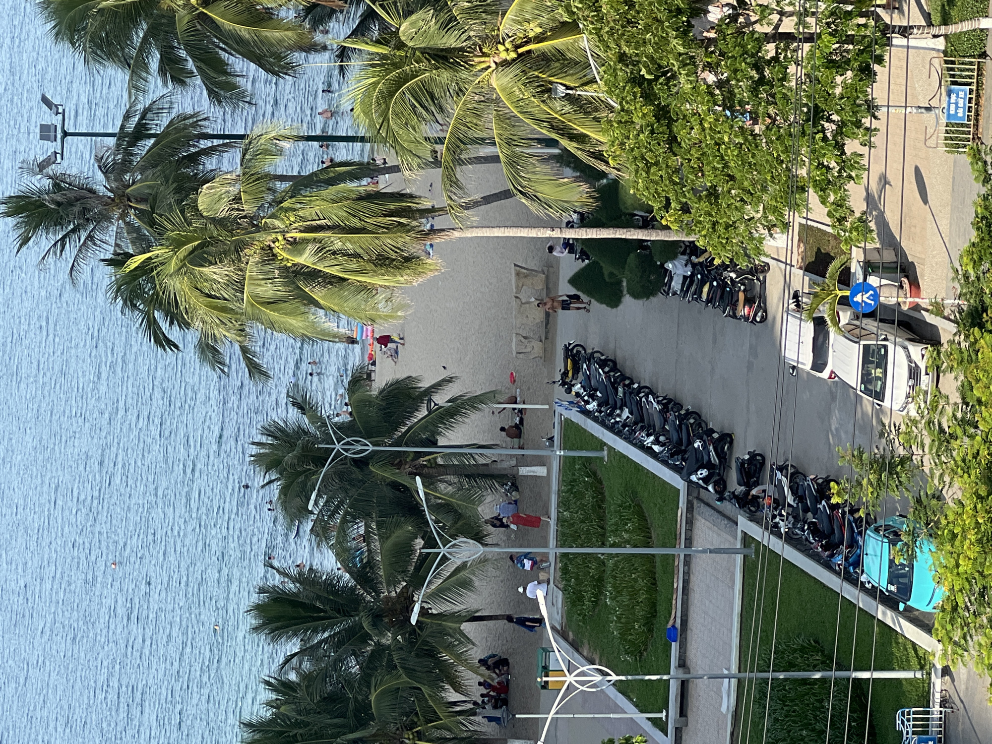 This aerial view captures a vibrant scene in Nha Trang, Vietnam, likely along its picturesque coastline. The turquoise ocean stretches towards the horizon, dotted with a few swimmers enjoying the water. A sandy beach lines the shore, populated by individuals and small groups relaxing or strolling. Lush palm trees, characteristic of tropical coastlines, are scattered along the beach and extend into the foreground, their fronds casting shadows.
In the mid-ground, a paved pathway separates the beach from a landscaped area featuring green grass and decorative plants. Along this pathway, a line of parked motorbikes suggests local transportation and access to the beach. Further down the road, a small, bright blue car is parked next to more motorbikes. A white van is also visible, parked on the opposite side of the road. A pedestrian crossing sign with a blue background and a white silhouette of a person walking is positioned near the parked vehicles.
The scene is one of leisurely activity, with a blend of natural beauty and urban elements. The presence of people enjoying the beach and ocean, coupled with the parked vehicles, indicates a popular public area. The lighting suggests it is daytime, with the sun casting distinct shadows.