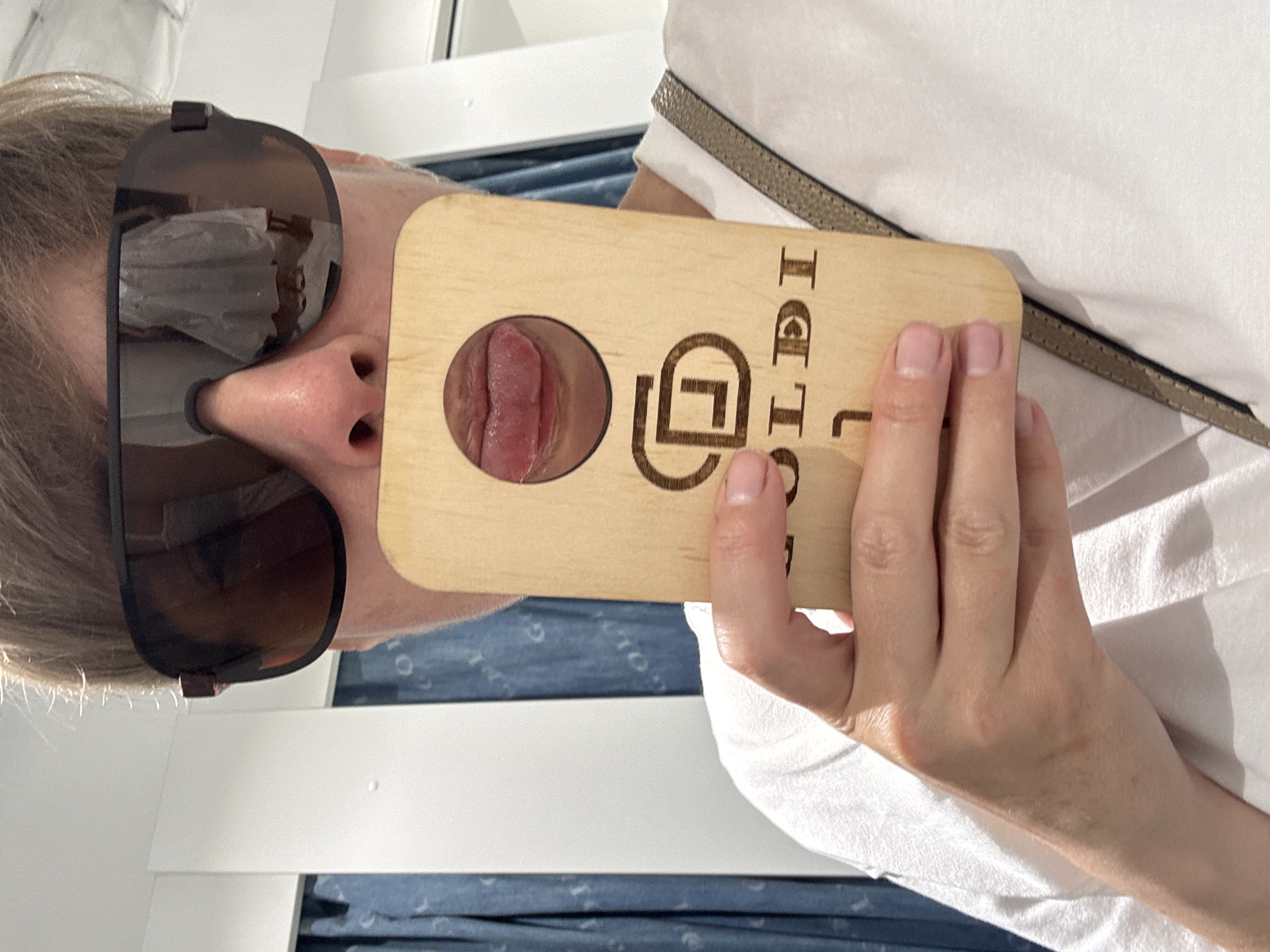 A person, seen from the chest up, is holding a wooden object with a circular cutout in front of their mouth. Their tongue is visible through the cutout, sticking out slightly. The person is wearing large, dark sunglasses and a white t-shirt. The wooden object has a logo and text etched into it. Behind the person, blue curtains are visible, and a white wall is to the left. The overall lighting suggests an indoor environment. The location is Unknown, Unknown.