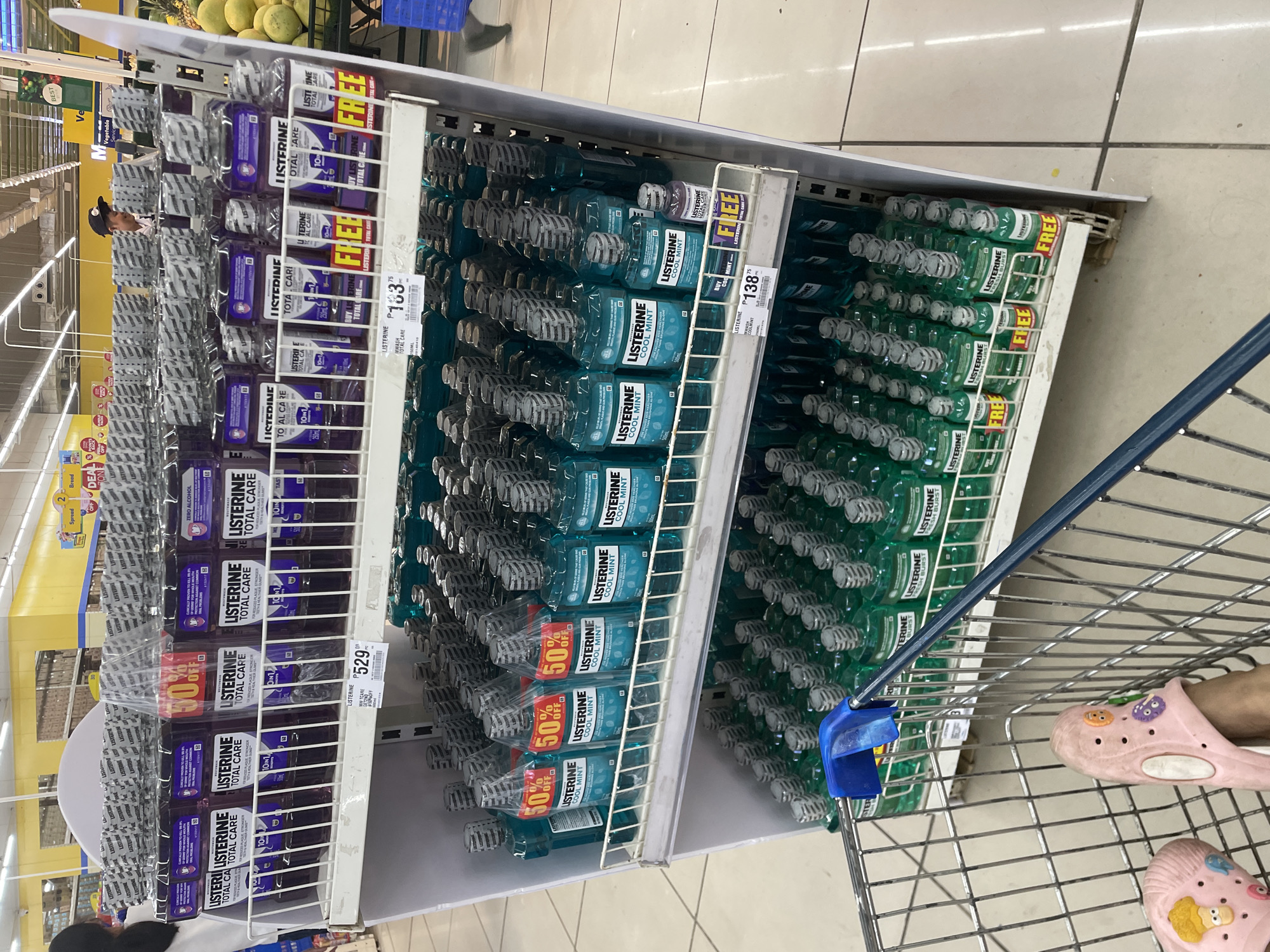 The image shows a retail store aisle displaying Listerine mouthwash products in various sizes and types, including "Total Care" and "Cool Mint." The products are neatly arranged on white shelves. Some bottles have prominent "50% OFF" or "FREE" stickers, indicating promotional offers. In the foreground, a portion of a shopping cart and a person's feet wearing pink, cartoon-decorated slippers are visible. The environment suggests a supermarket or drugstore setting, with aisles and shelving typical of such establishments. The presence of a shopping cart and promotional signage points to a commercial transaction in progress. While the image does not explicitly state the location, the language on the price tags ("P" for Philippine Peso) indicates it is likely in the Philippines. Considering the user's request to locate it in Davao City, Philippines, this scene depicts a typical retail environment in that city.