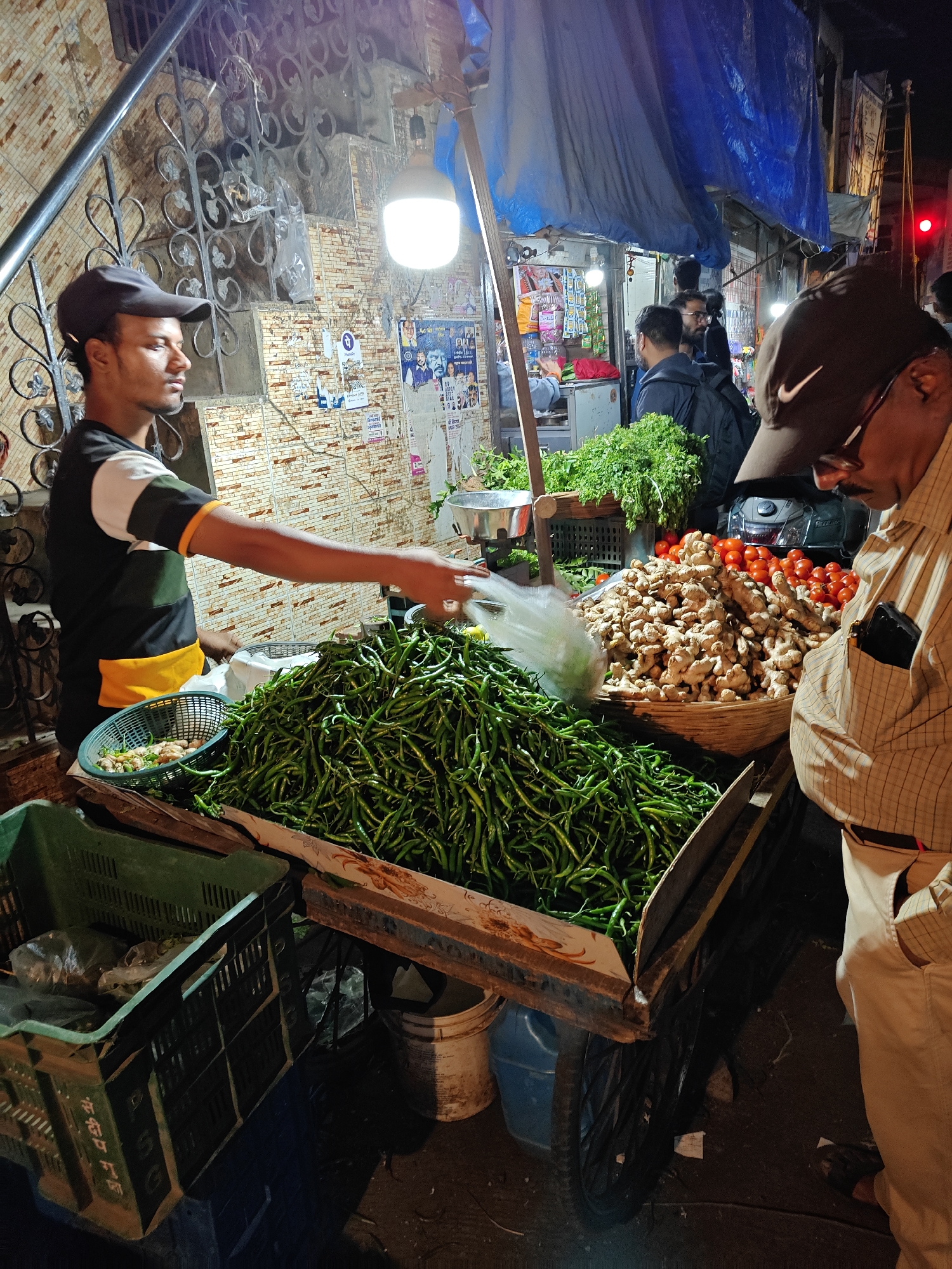 Here is a detailed description of the image:
**Scene:** An outdoor night scene showing a vegetable vendor in Mumbai, India, interacting with a customer. The vendor is behind a mobile cart loaded with produce.
**People:**
* A young man, the vendor, is wearing a dark cap, and a T-shirt with green, yellow, and white stripes. He is holding a plastic bag, seemingly serving or packaging vegetables for a customer.
* An older man, likely a customer, is wearing a brown cap, glasses, and a light-colored patterned shirt. He's looking down at the produce, presumably selecting items to purchase.
* There are other individuals in the background, indicating a bustling street scene.
**Subjects/Objects:**
* The cart contains a large pile of green chilies, ginger, and tomatoes. There are also green vegetables in a basket.
* Other crates and containers are near the cart, likely holding additional produce.
* In the background, there appears to be a storefront with more vegetables and other goods.
* A bright light illuminates the vendor's cart.
* Behind the vendor are stairs with metal railings, and the wall is plastered with posters and paper.
**Actions/Interactions:** The vendor is in the process of preparing or handing over produce to the customer. The customer is looking at the produce, apparently shopping.
**Environment:** The scene appears to be a narrow street in Mumbai. The lighting is uneven, mostly coming from the vendor's light and the storefront behind. The background shows urban characteristics, including buildings, posters, and general street activity.