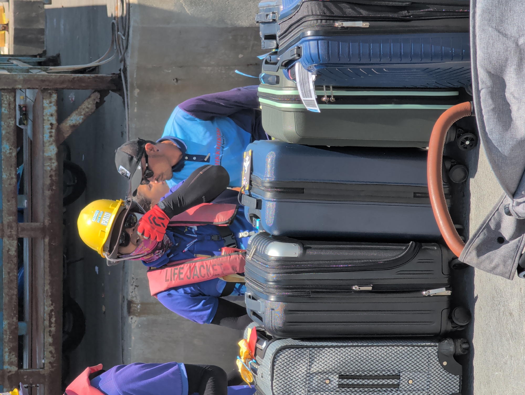 Two individuals are present in the scene. One individual is wearing a yellow hard hat with stickers, red gloves, and a life jacket. The other individual is wearing a baseball cap, sunglasses, and also a life jacket. The individuals are next to a stack of luggage. The luggage includes suitcases of various sizes and colors. The scene is located in ตำบลเกาะพะงัน, Thailand, adjacent to a metal structure with wheels.