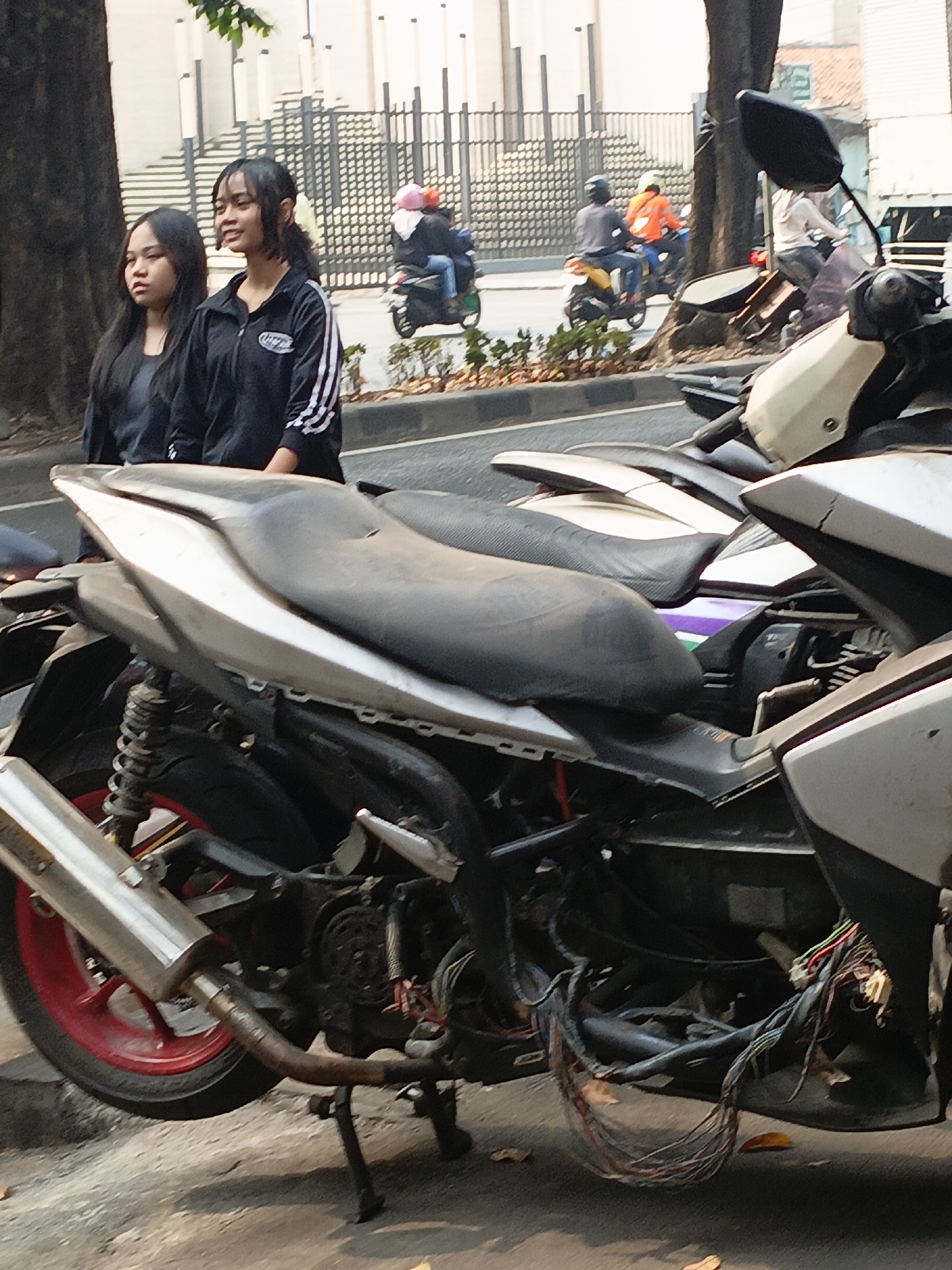 Two young Indonesian women stand near a street in East Jakarta. The woman on the left has long, dark hair and wears a dark shirt. The woman on the right has dark hair with bangs and wears a black jacket with white stripes on the sleeves. Several motorcycles are parked on the street, with one partially in the foreground, showing its exposed engine and wiring. In the background, other motorcycles are visible, some with riders. The scene takes place outdoors, with trees and a fence lining the street.