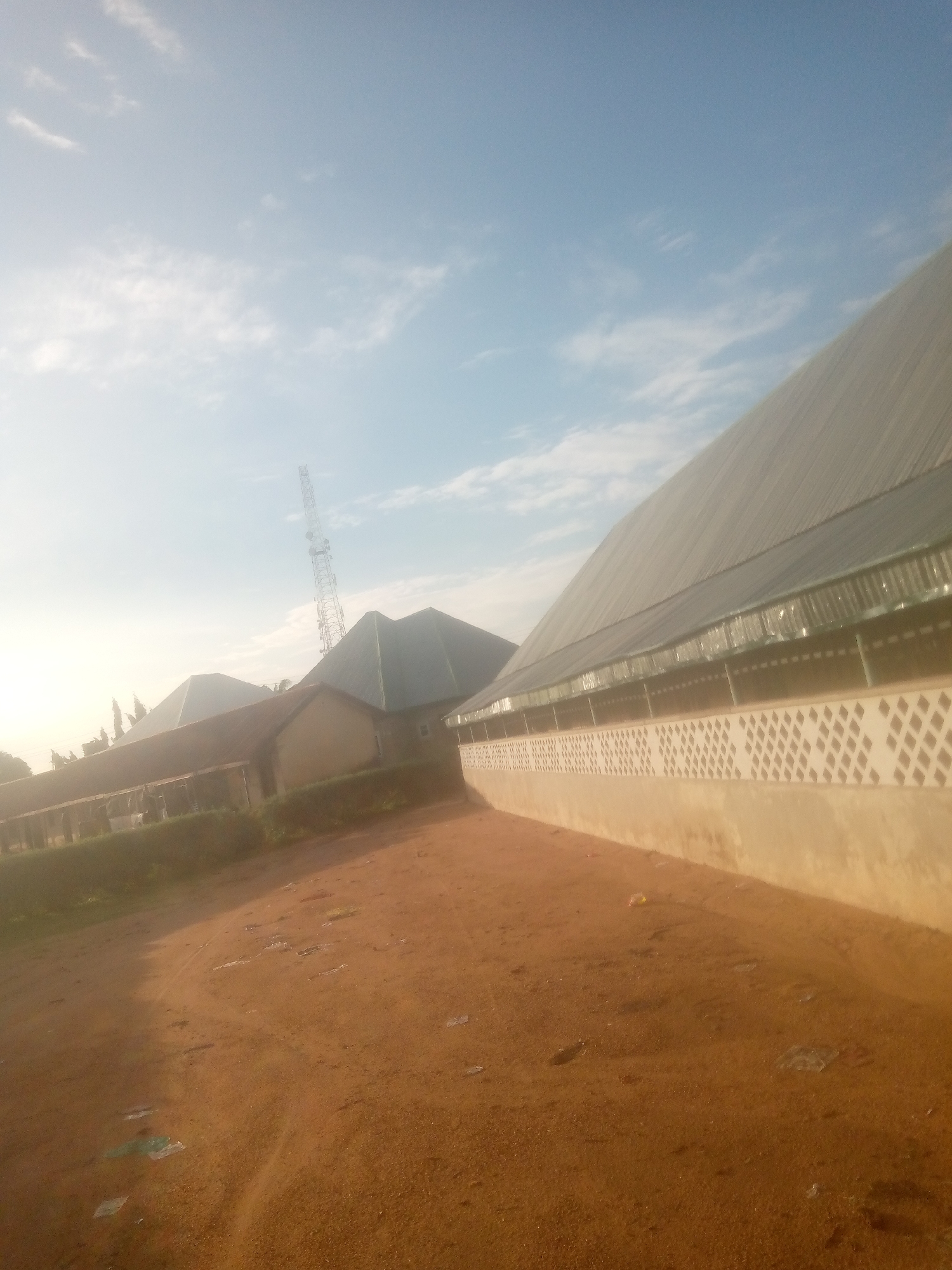 This image, taken in Jalingo, Nigeria, displays an outdoor scene focused on buildings and a reddish-brown unpaved ground under a bright, partially clouded sky. In the foreground, an uneven reddish-brown dirt surface is visible, strewn with scattered pieces of clear plastic debris. To the right, a large structure with a steeply pitched, dark gray corrugated metal roof extends diagonally across the frame. Its lower wall features a white diamond-patterned lattice design, likely for ventilation, set above a solid beige base. In the mid-ground, to the left, several other buildings are present; one has a dark green hipped roof, and another a flatter reddish-brown corrugated roof, partially obscured by a strong atmospheric haze. Behind these structures, a tall, silvery-gray lattice communication tower rises prominently against the bright sky. A narrow strip of green vegetation borders the unpaved ground along the base of the left-side buildings. No individuals are present, and no actions are depicted. The lighting indicates strong sunlight, casting a significant glare or haze on the left side of the frame, characteristic of either early morning or late afternoon.