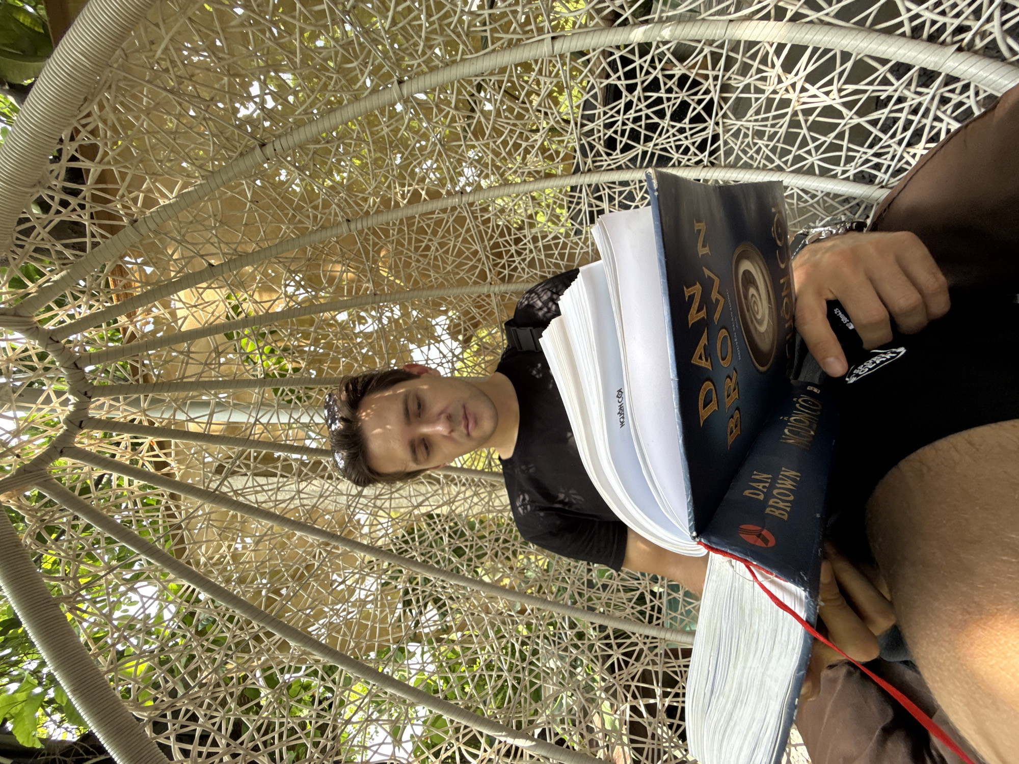 A man is depicted relaxing and reading outdoors in a brightly lit setting, likely in Diên Khánh, Vietnam. He is seated in a distinctive white, intricately woven or netted chair, possibly an egg chair, which provides partial shade and allows glimpses of green foliage and bright sky through its pattern. The photograph is taken from a low angle, looking upwards at the man, giving the image a unique perspective.
The man, appearing to be in his 20s or 30s, has short dark hair and wears sunglasses pushed up onto his head. He is dressed in a dark t-shirt, with the word "SEKAI" partially visible on the fabric. His eyes are closed or heavily lidded, suggesting a moment of deep concentration, relaxation, or perhaps light dozing while holding a large, blue hardcover book open.
The book is prominently open in his hands, with a red ribbon bookmark dangling from the bottom. The cover clearly shows "DAN BROWN" as the author, and the title appears to be "ORIGIN," featuring a stylized spiral or coffee bean-like symbol. In his right hand, the man holds a black pen or marker, which has "SOMA-25" written on its side. A metallic watch is visible on his left wrist. The strong sunlight and dappled shadows indicate it is daytime, likely midday or afternoon, suggesting pleasant, sunny weather. The overall scene conveys a sense of peaceful leisure and immersion in reading.