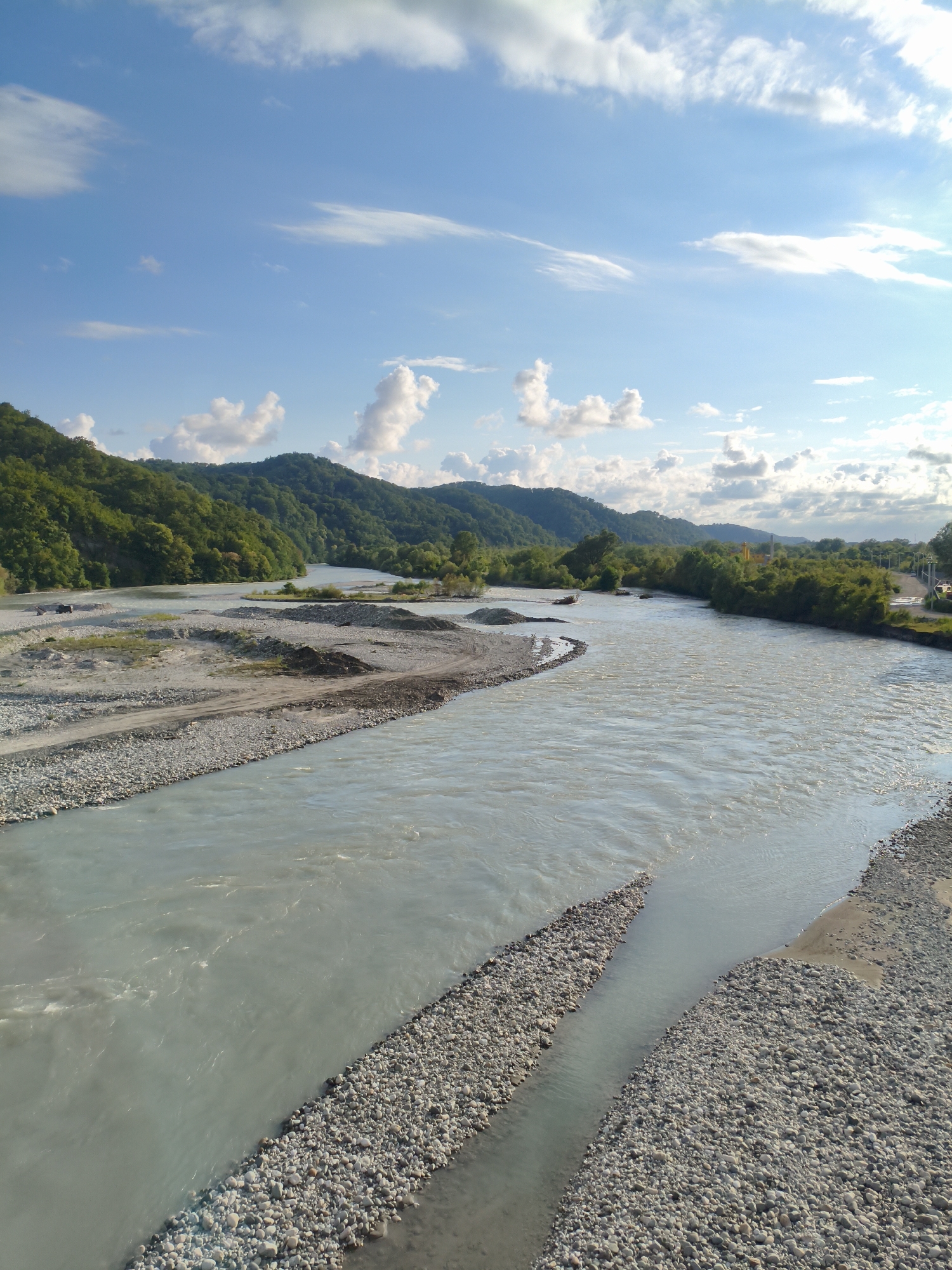 The image captures a wide, slow-moving river flowing through a valley. The riverbed is lined with grey and white pebbles, with some areas of sand and sediment visible. Lush green trees cover the rolling hills that flank the river on both sides. The sky above is a bright blue, dotted with fluffy white clouds, suggesting a pleasant day.
The scene appears to be a natural landscape, possibly in a rural or mountainous region. The presence of a road and some structures in the distance, particularly on the right side of the image, hints at some human presence or development in the area, but the overall impression is one of natural beauty. There are no people or specific objects that draw the viewer's attention, allowing the focus to remain on the grandeur of the landscape. The time of day appears to be either late morning or early afternoon, given the bright sunlight and the shadows cast. The weather seems fair and clear. The location context provided, Tsaniguarta, Abkhazia, places this scene within a specific geographical area known for its scenic beauty. The image conveys a sense of tranquility and the raw power of nature.