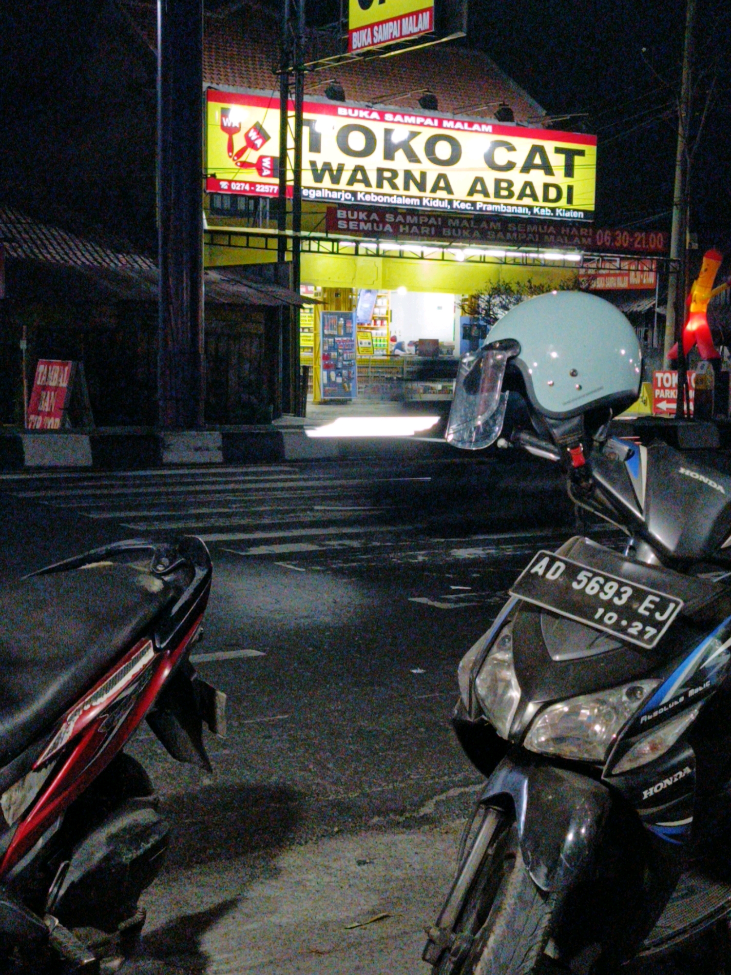The image depicts a nighttime street scene in Kebondalem Kidul, Indonesia, featuring a parked motorcycle in the foreground and a brightly lit shop in the background. The motorcycle, partially visible, is black with red accents and has a light blue helmet resting on its handlebars. A license plate is attached to the front of the motorcycle, reading "AD 5693 EJ 10.27".
Behind the motorcycle, a shop identified by a prominent yellow and red sign reads "TOKO CAT WARNA ABADI". Below this, additional text indicates the shop is located in "Kebondalem Kidul, Kec. Prambanan, Kab. Klaten". Further text on the shop's signage suggests it is open "BUKA SAMPAI MALAM" (Open until night) and "SETIAP HARI" (Every day) with operating hours from "06:30-21:00". The shop's interior is visible through illuminated windows, displaying shelves stocked with various products. A red A-frame sign that reads "TAMBAL BAN" (Tire Repair) is positioned on the sidewalk to the left of the shop. The street is paved and appears to be a crosswalk with striped markings. The overall atmosphere is illuminated by artificial lights from the shop and streetlights.