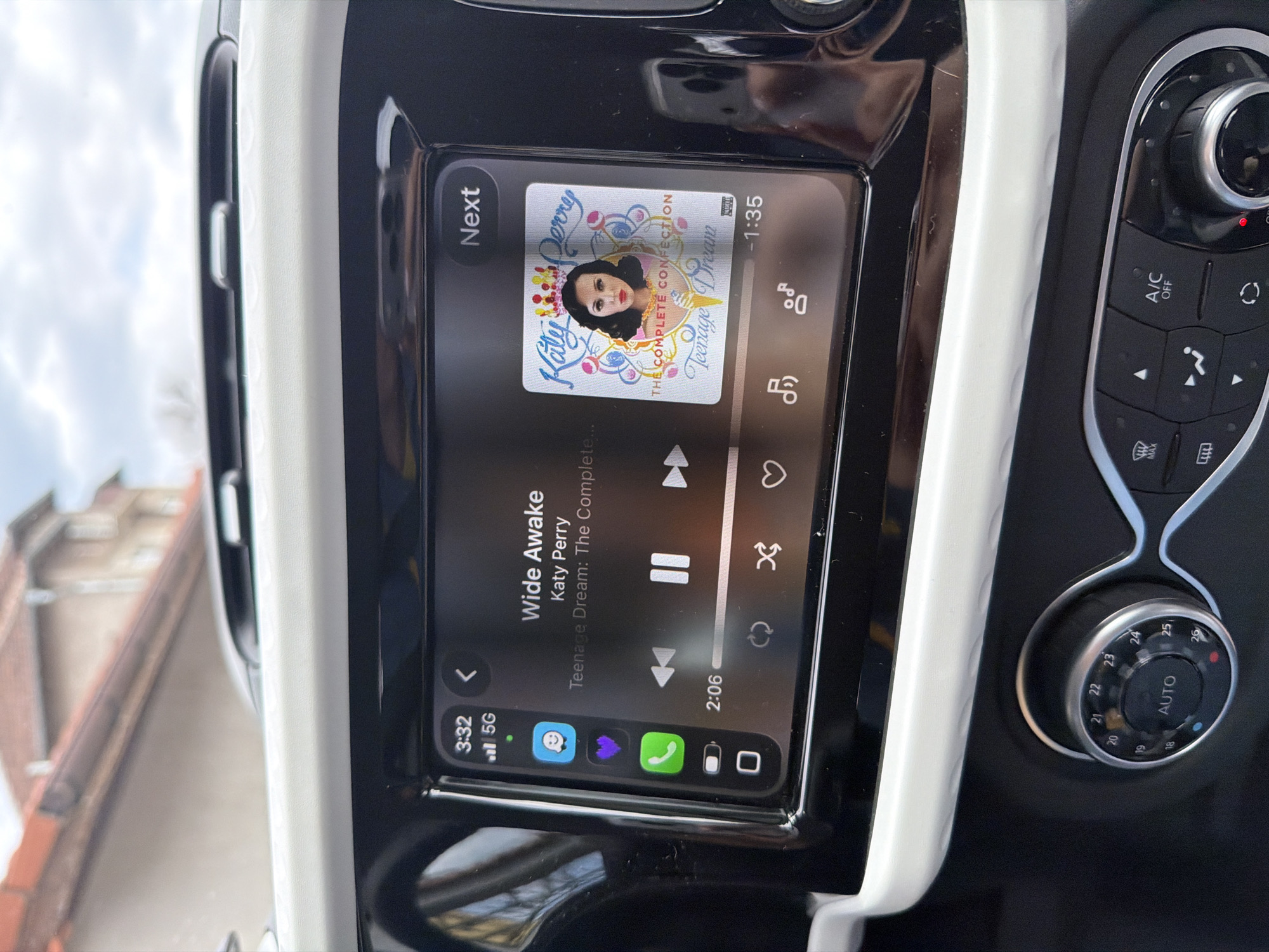 The image shows the in-car entertainment system of a vehicle. The system displays the song "Wide Awake" by Katy Perry from the album "Teenage Dream: The Complete Confection." The track is at the 2:06 mark, with 1:35 remaining. The system is connected to a phone, as evidenced by the icons on the bottom left showing connection to 5G and phone applications. To the right of the screen are vehicle controls for the air conditioning, including temperature and circulation settings. The environment outside the car suggests the image may have been taken in Buhovo, Bulgaria due to the presence of a building with a red tile roof. No people are directly visible.