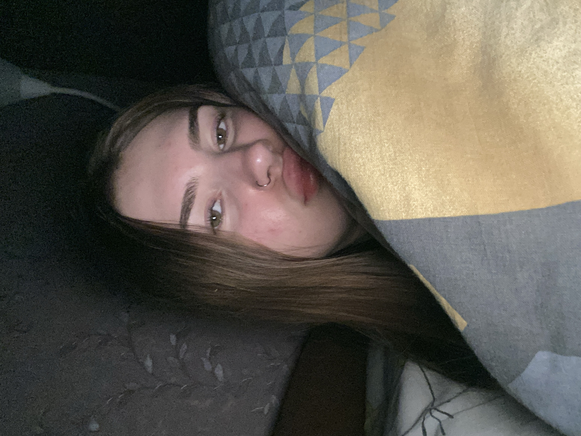 A young female with long, brown hair and brown eyes is seen in a close-up portrait. She is wearing a silver nose ring. Her lips are slightly pursed, and she is looking directly at the camera. She is partially covered by a blanket with a geometric pattern of grey, yellow, and blue triangles. The background is dark and appears to be indoors, possibly in a bedroom. The lighting suggests it might be nighttime or a dimly lit room. The environment appears to be the interior of a dwelling in the city of Dyakovo, Russia.