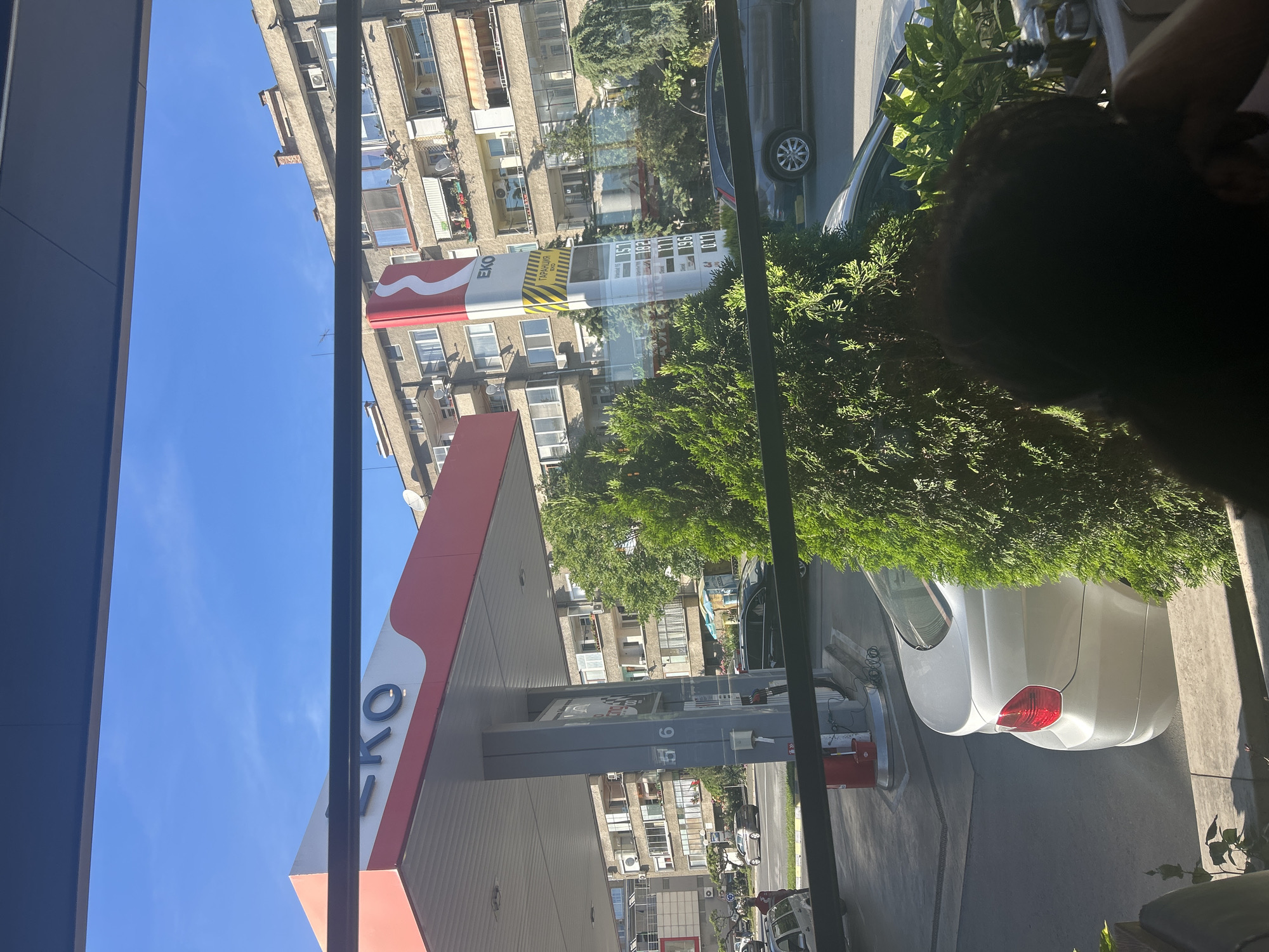 The image shows a view from inside a building, looking out at an EKO gas station in Kazanlak, Bulgaria. The gas station has a red and white canopy with the "EKO" logo. Behind the gas station are several apartment buildings. There are cars parked at the gas pumps. The sky is blue. In the foreground, there is a portion of a person's head and a plant.