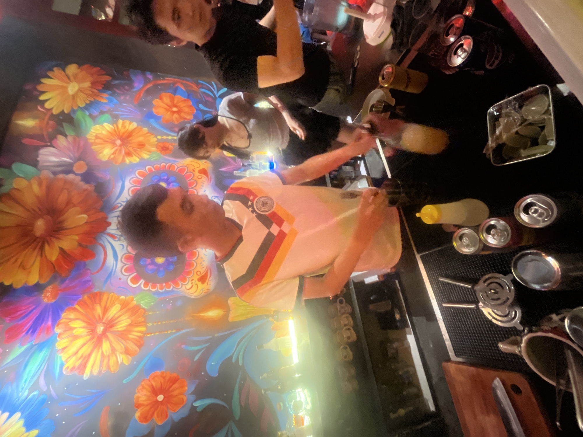 A group of people are behind a bar with a vibrant mural of flowers and a candle on the wall. A man in a white t-shirt with red, black, and yellow stripes on the collar and chest area is preparing a drink, holding a bottle and a strainer. To his right, a woman in a grey tank top and black shorts is also present. To the far right, another man in a black t-shirt is working. The bar counter is stocked with various items, including bottles, cans, strainers, and a small metal container with lime slices. The lighting is dim, with bright accents from the bottles on the shelf behind the bar. The overall setting appears to be a bar or lounge.