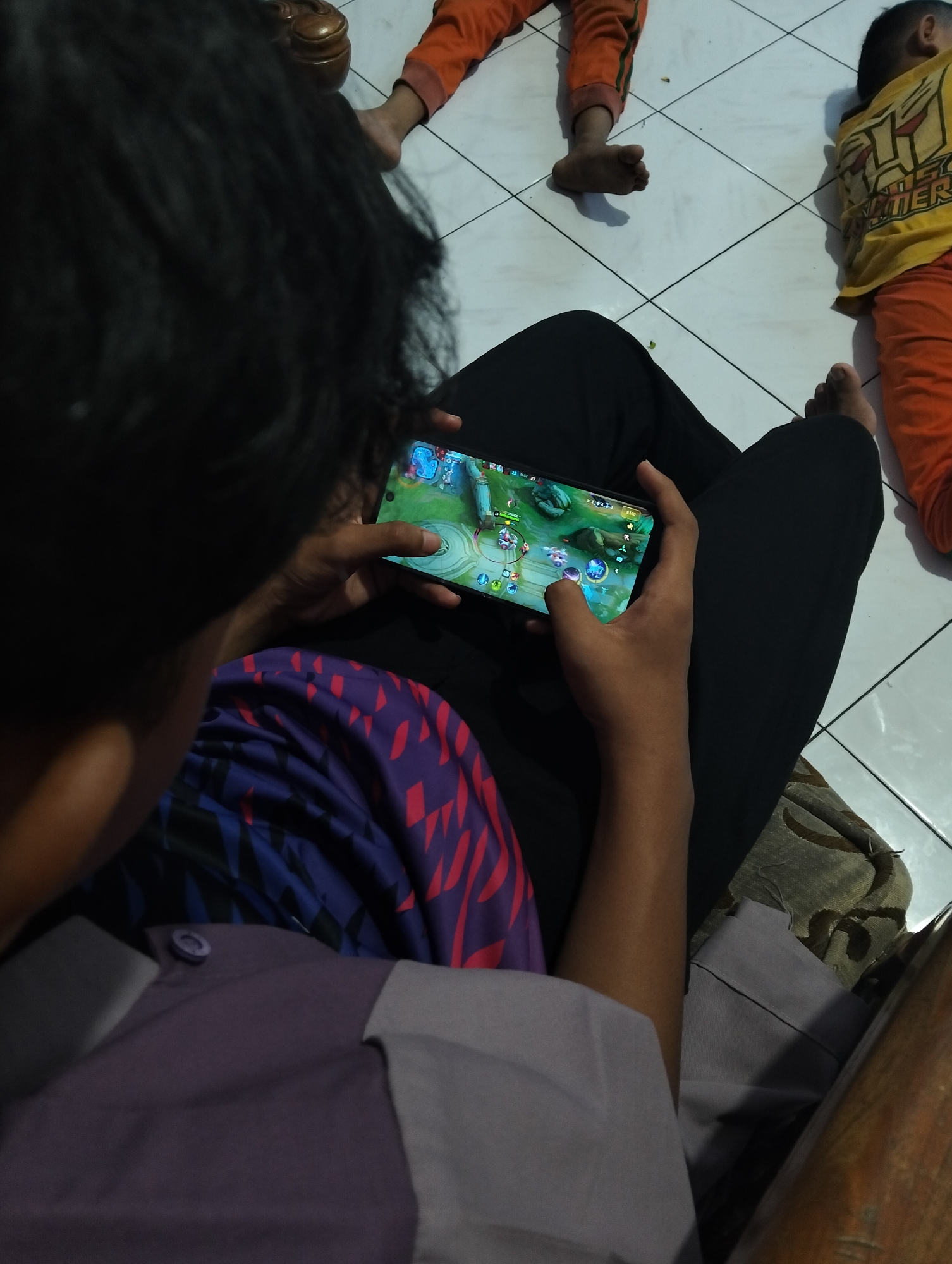 In a domestic setting in the City of Medan, Indonesia, a person is seated, engrossed in playing a mobile game on their smartphone. The phone displays a colorful game interface, with the player's fingers actively interacting with the screen. The individual is wearing black pants and a patterned shirt in shades of purple and blue. In the background, two children are lying on the floor, one wearing an orange shirt and pants, and the other a yellow shirt with an orange undershirt. The floor is tiled with a white grid pattern. The lighting suggests an indoor environment, possibly in the late afternoon or evening.