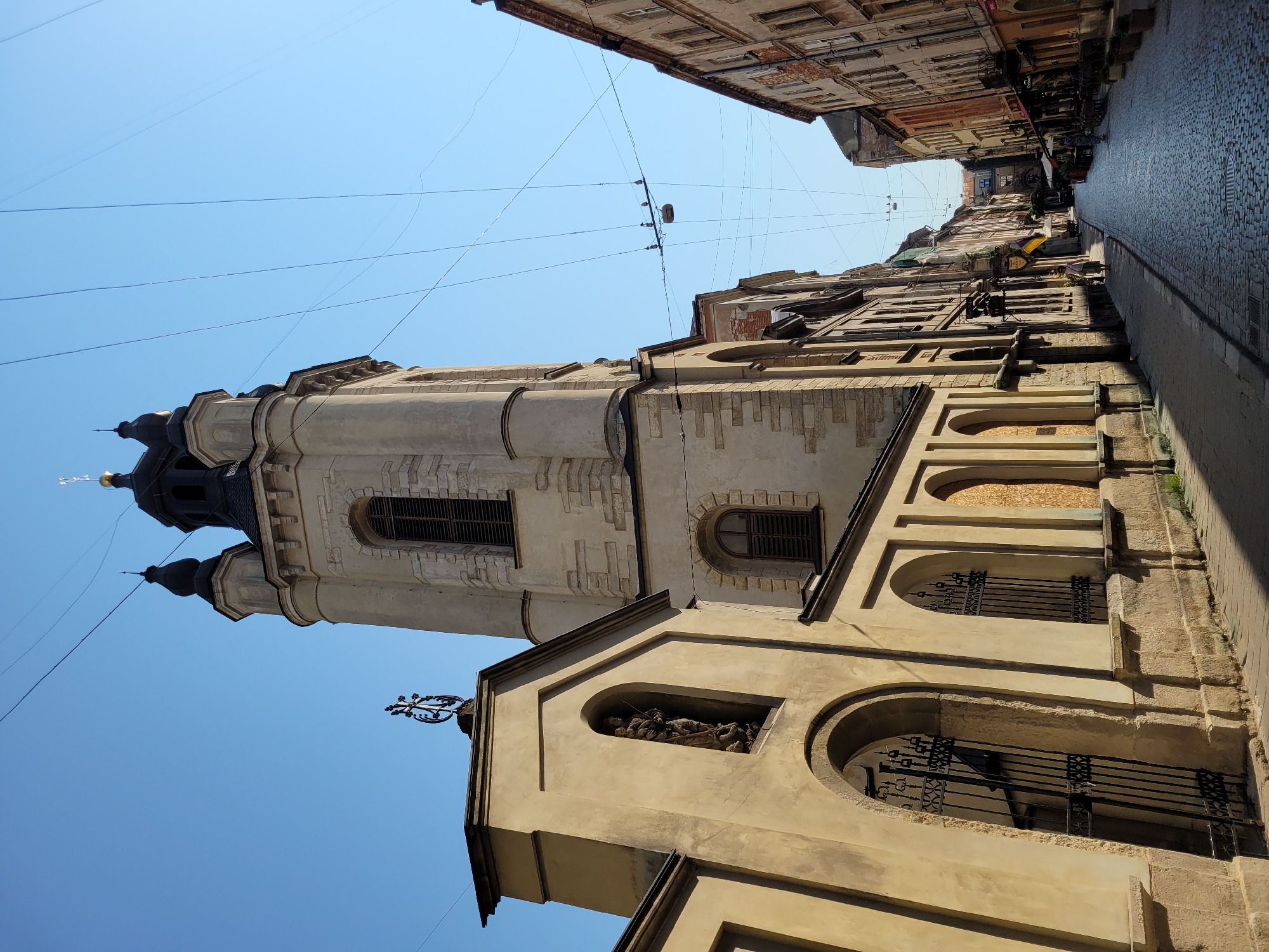 The photograph captures a sunlit street scene in an unknown city, likely in Europe, with a clear blue sky overhead. The perspective is low, looking up a cobblestone street lined with historic buildings. On the left, a pale yellow stucco building features a prominent arched entryway with black wrought-iron gates, leading to a dark interior. Above this entrance, an arched niche contains a religious statue. Adjacent to this, a tall, light-stone bell tower with a dark, multi-tiered roof rises towards the sky. The tower has arched windows and stone block detailing. The street itself is paved with cobblestones and is relatively narrow, with an inviting perspective drawing the viewer down its length. Along the right side of the street, older, multi-story buildings with worn facades and balconies line the thoroughfare, their colors ranging from muted oranges to grays. A few people are visible in the distance, walking along the street. Overhead, a network of dark wires crisscrosses the sky, some supporting a lamppost, suggesting the presence of electrical infrastructure. The lighting suggests it is daytime, with strong shadows cast by the buildings, indicating a bright sun.