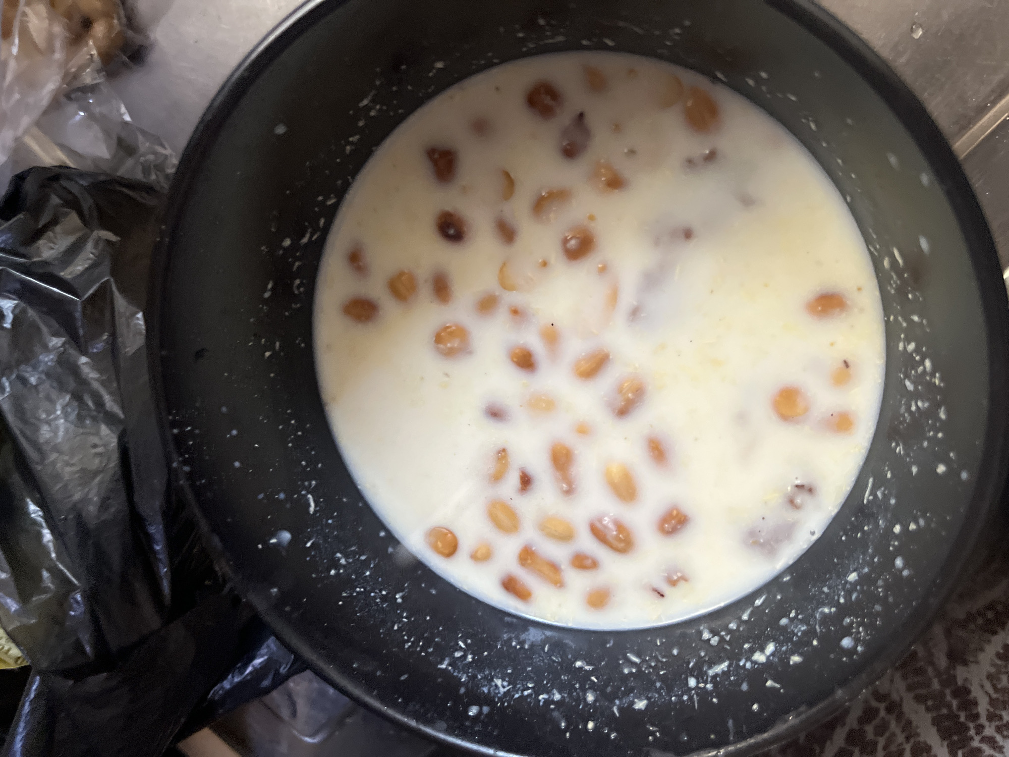 A close-up overhead shot shows a black bowl filled with a creamy white liquid and small, light brown pieces scattered throughout. The pieces appear to be some sort of grain or small nuts, possibly raisins or peanuts, floating in milk or a similar dairy-based liquid. The bowl itself has a textured, slightly gritty surface on the rim and the surrounding area, with some white splatters visible on the black exterior. To the left of the bowl, a crumpled black plastic bag is visible, partially obscuring a metallic surface and some indistinct objects wrapped in clear plastic. The overall scene suggests a food preparation or serving context, possibly in a kitchen or domestic setting, given the presence of the plastic bag and the informal presentation of the food. The lighting appears to be artificial, and there are no clear indicators of time of day or weather. The setting appears to be indoors. No visible text is present in the image.