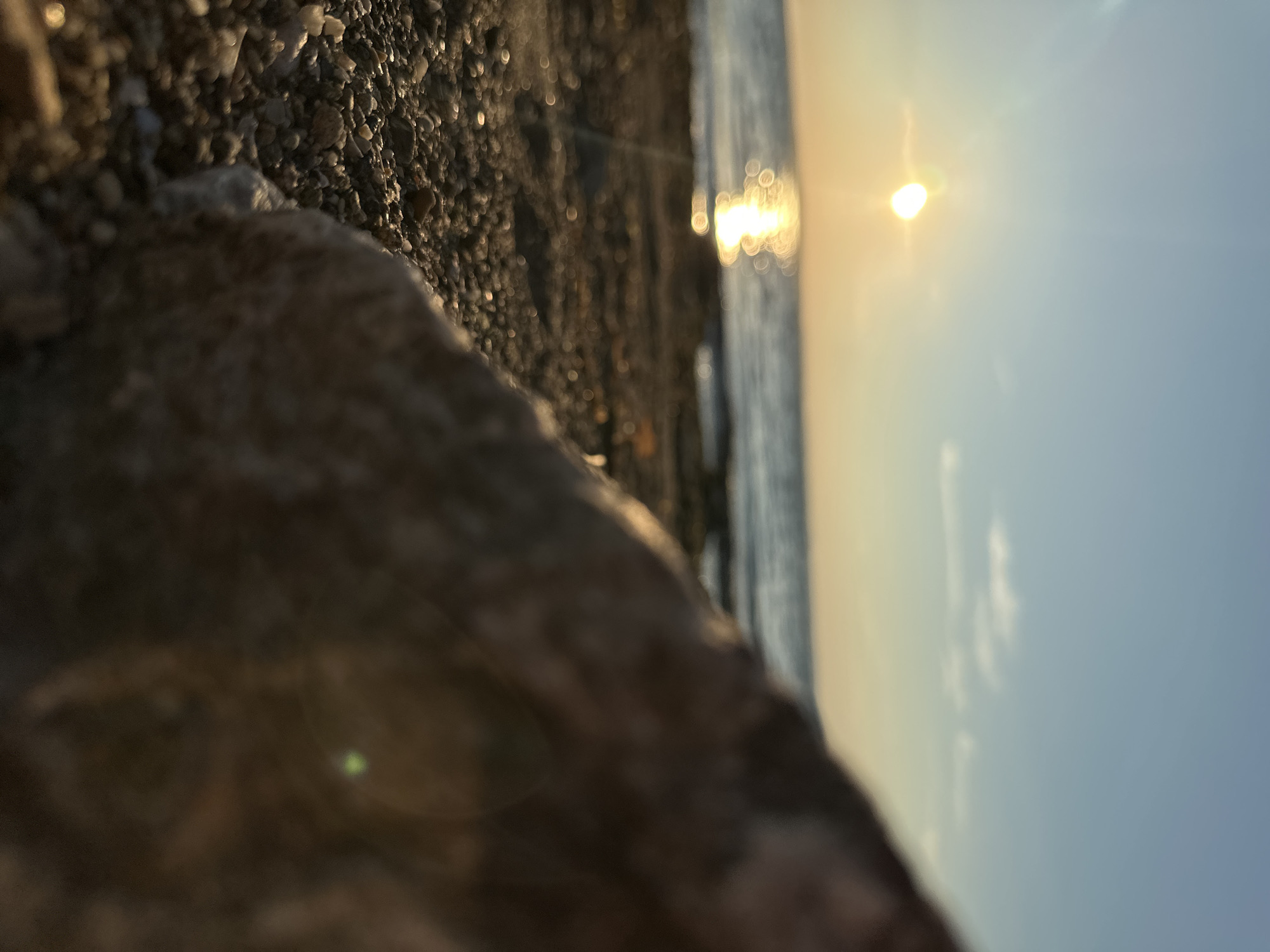 A low-angle, close-up shot of a beach in Energetik, Russia, with the sun setting over the Sea of Azov. In the foreground, a large rock is partially in focus, with the ground covered in pebbles and sand. The sun is low on the horizon, casting a golden reflection on the water. The sky is a clear blue with a few wispy clouds. The image appears to have been taken during the golden hour.