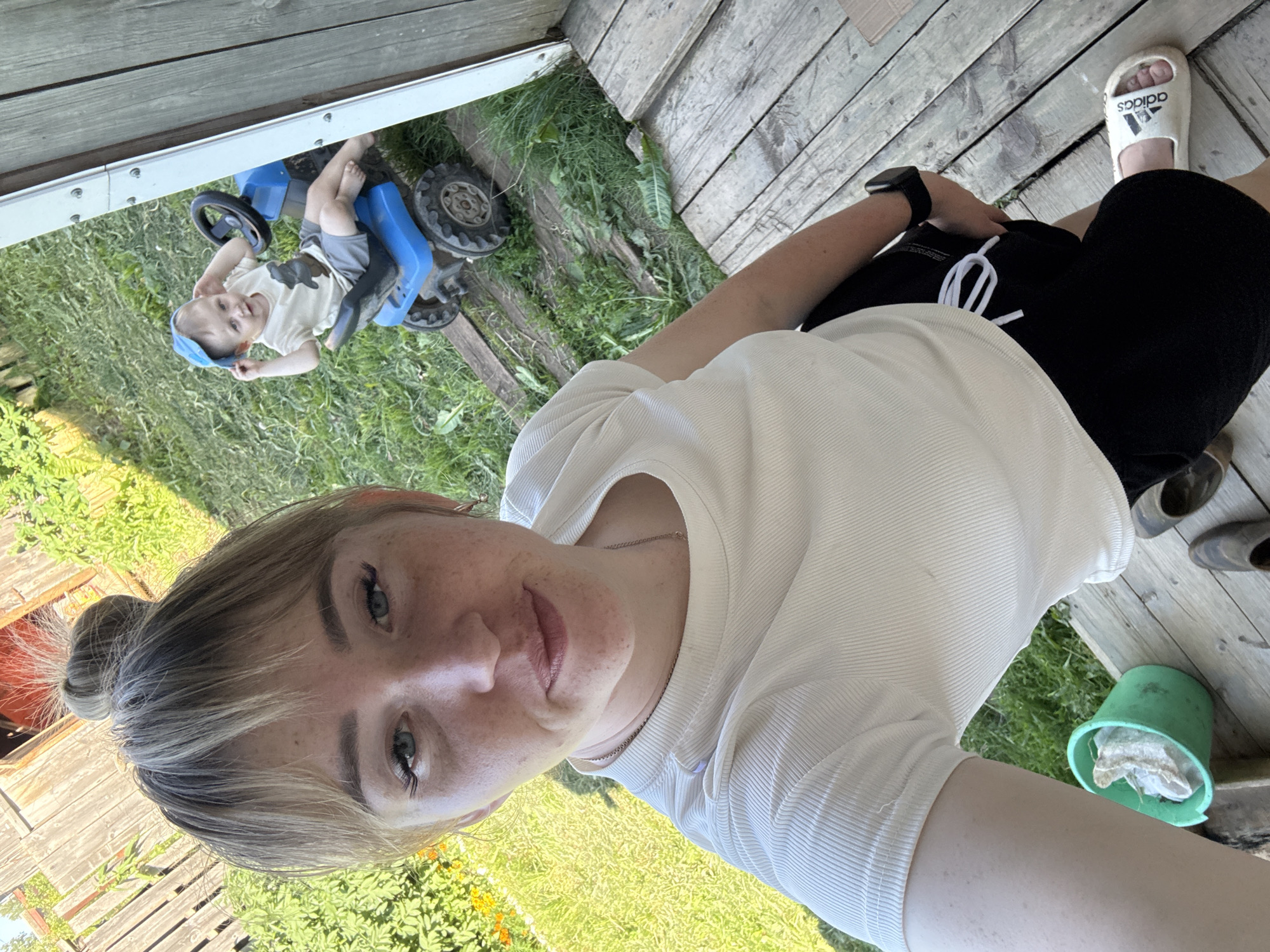 A woman with blonde hair pulled into a bun and freckles on her face is taking a selfie outdoors. She is wearing a white ribbed t-shirt and black shorts. To her right, a young child wearing a beige t-shirt, grey shorts, and a blue cap is sitting on a blue toy tractor. The background consists of a wooden fence, green grass, and a wooden porch. The setting appears to be a backyard in a rural area. The image was taken on a sunny day.