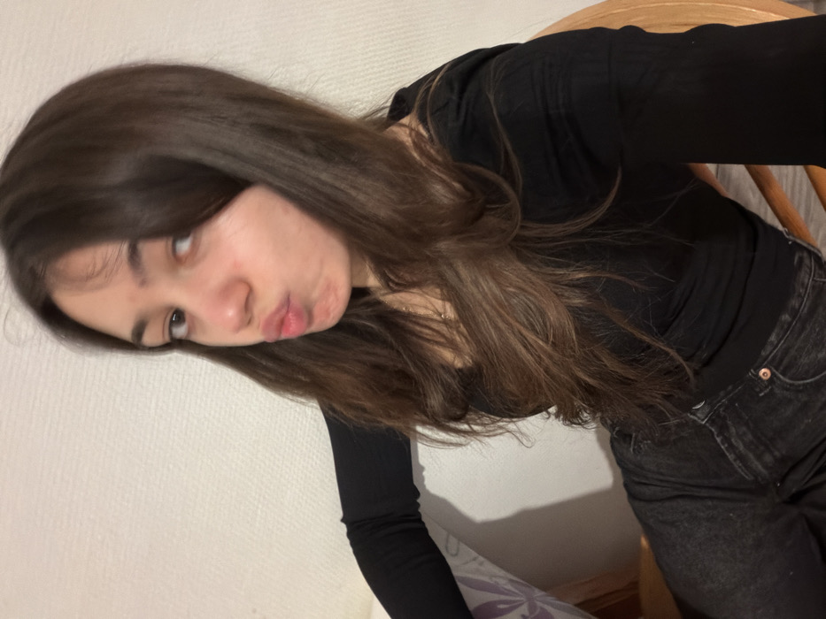 A young woman with long, brown hair is looking towards the camera while puckering her lips. She's wearing a black long-sleeved shirt and dark denim jeans. She appears to be sitting on a wooden chair, and the background is a plain, textured white wall. The image is slightly tilted, giving a casual, informal impression.